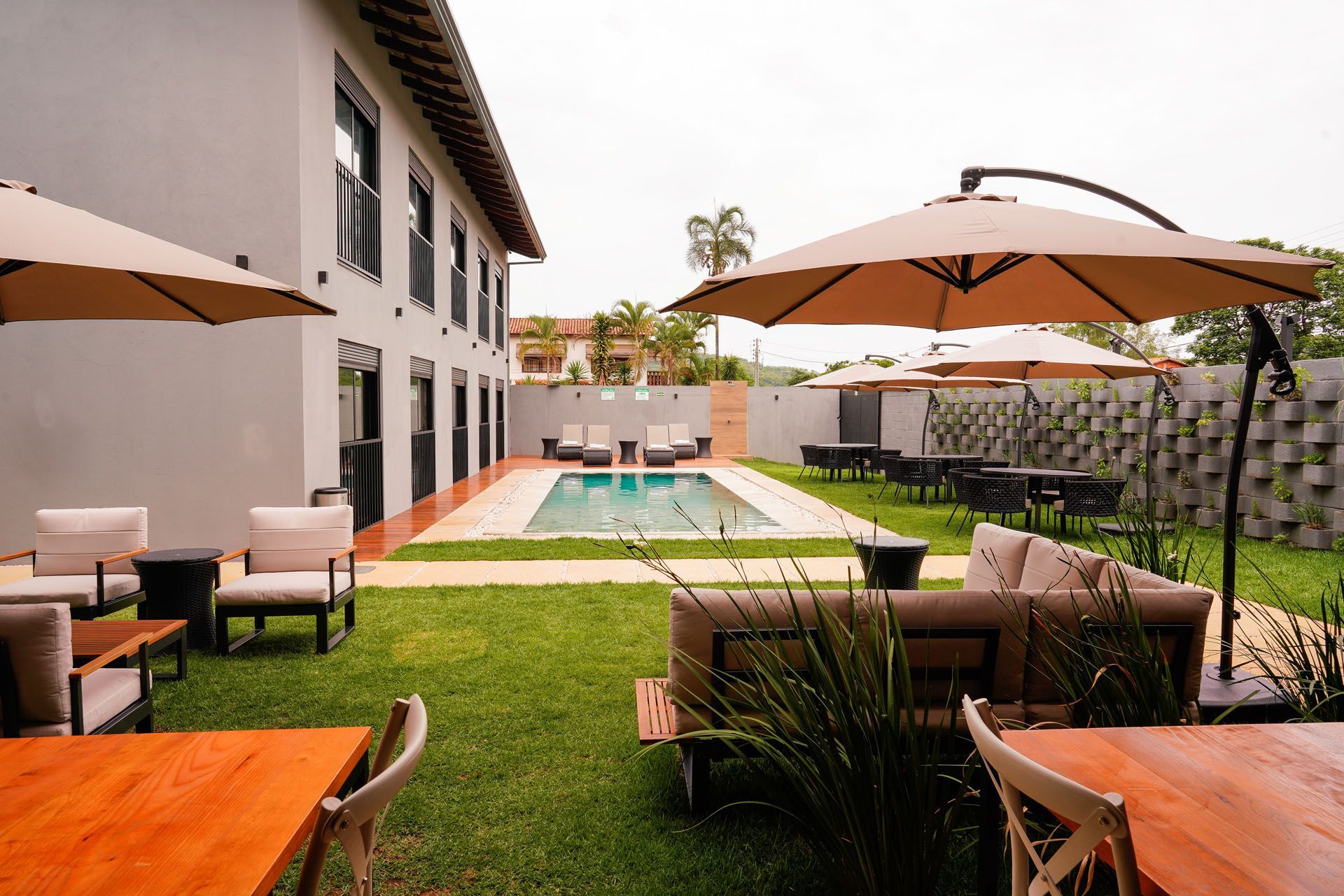Pátio externo com piscina, guarda-sóis, mesas e espreguiçadeiras ao lado de um prédio de dois andares em um dia nublado.
