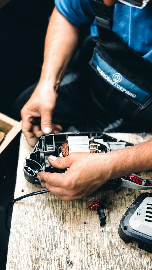 Technician repairing a disassembled power tool on a worn workbench with tools nearby