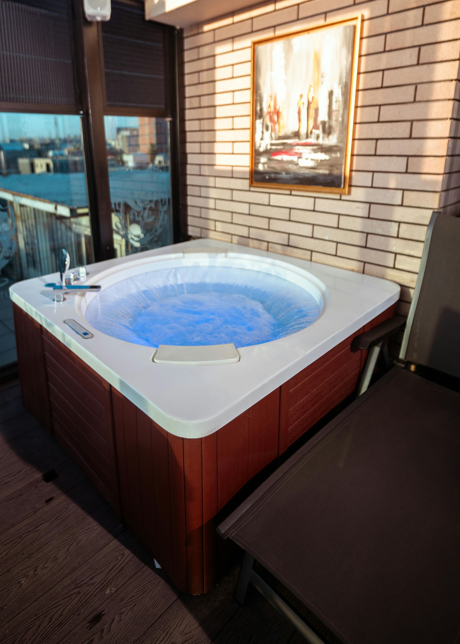 Wooden hot tub on a patio beside a brick wall and framed artwork, with blue water bubbling inside.