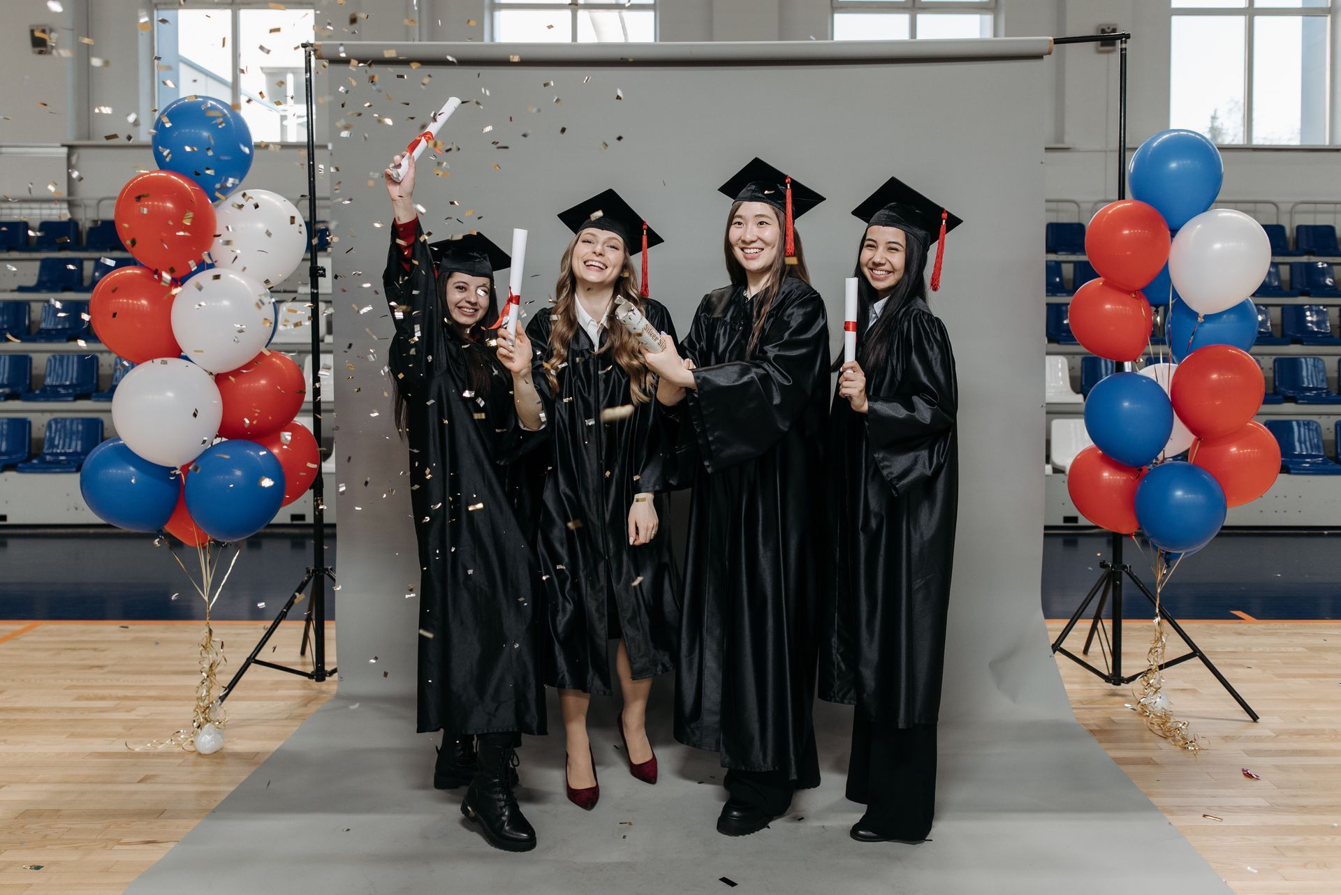 Four graduates in caps and gowns celebrate with confetti between red, white, and blue balloon columns.