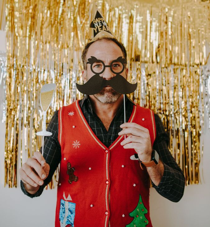 Man in red vest and festive glasses holding props in front of gold tinsel backdrop