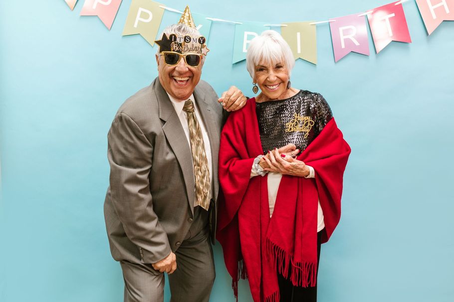 Two people in festive hats posing in front of a “HAPPY BIRTHDAY” banner, one holding a red shawl.