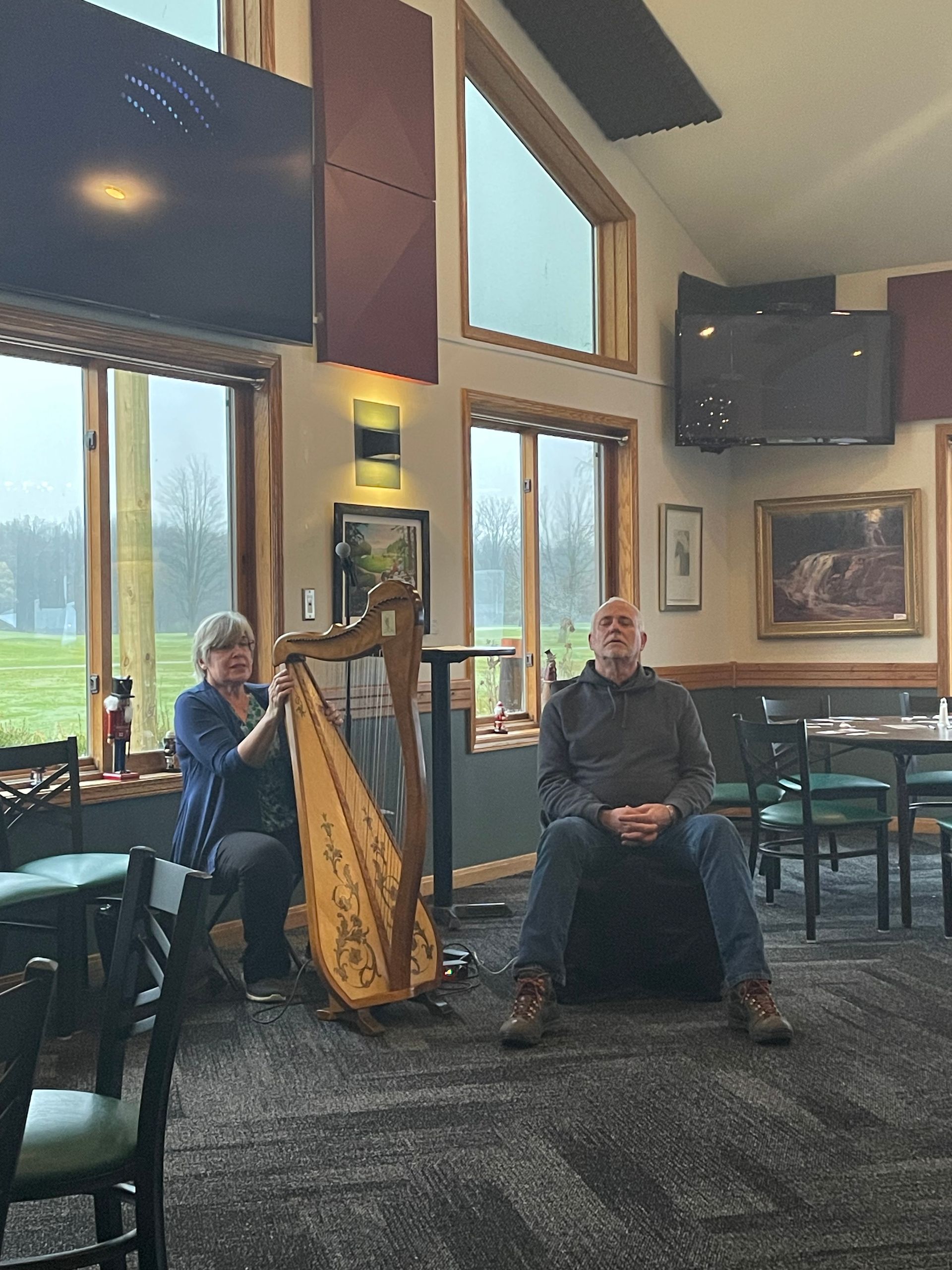 A woman is playing a harp while a man sits in a chair.