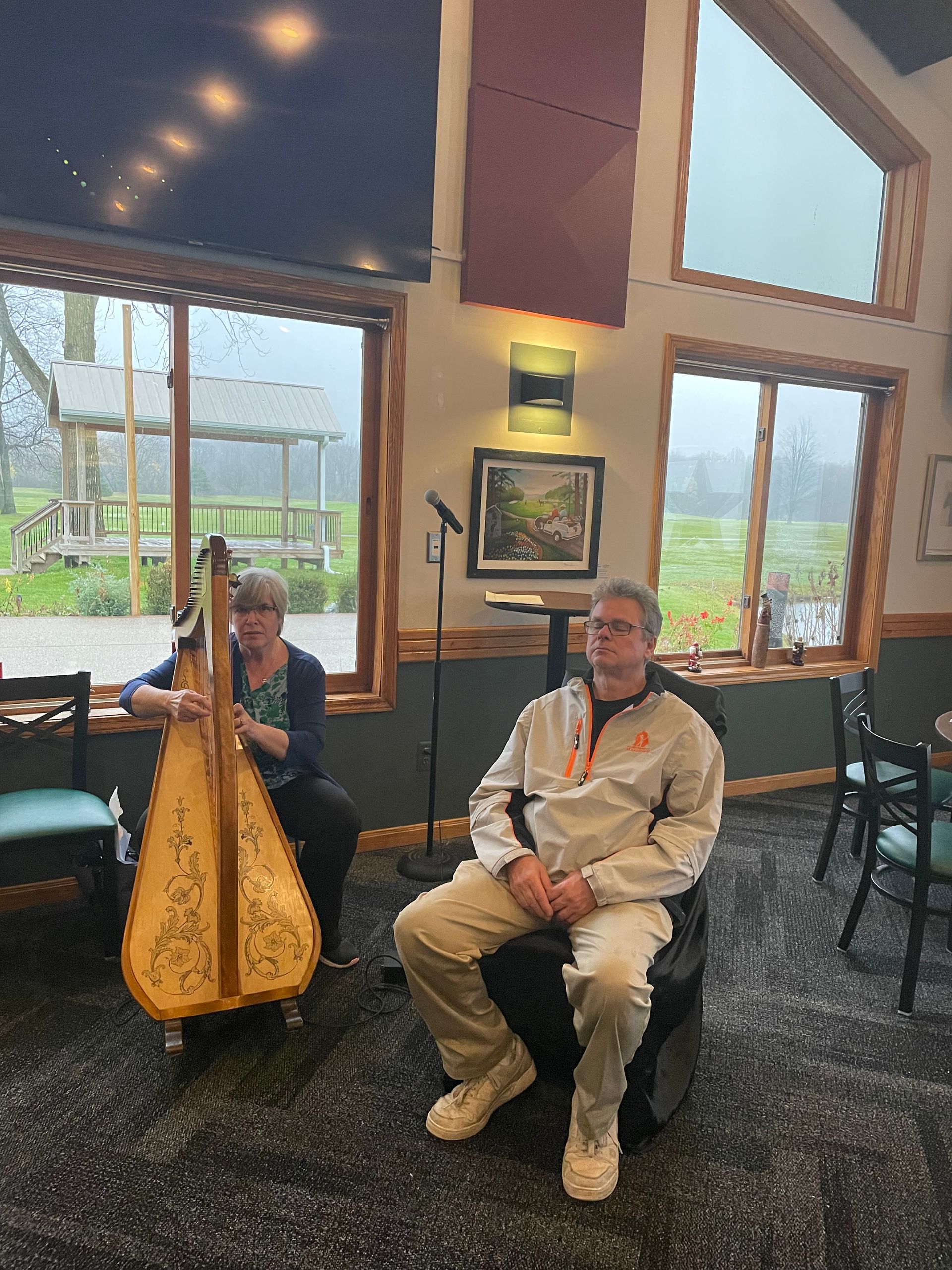 A woman is playing a harp and a man is sitting in a chair.