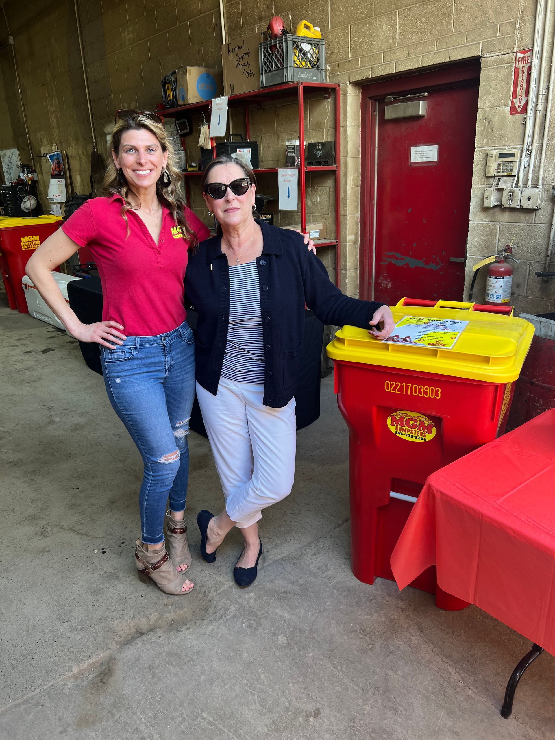 Two women standing next to each other in front of a yellow trash can that says 'recycle' on it