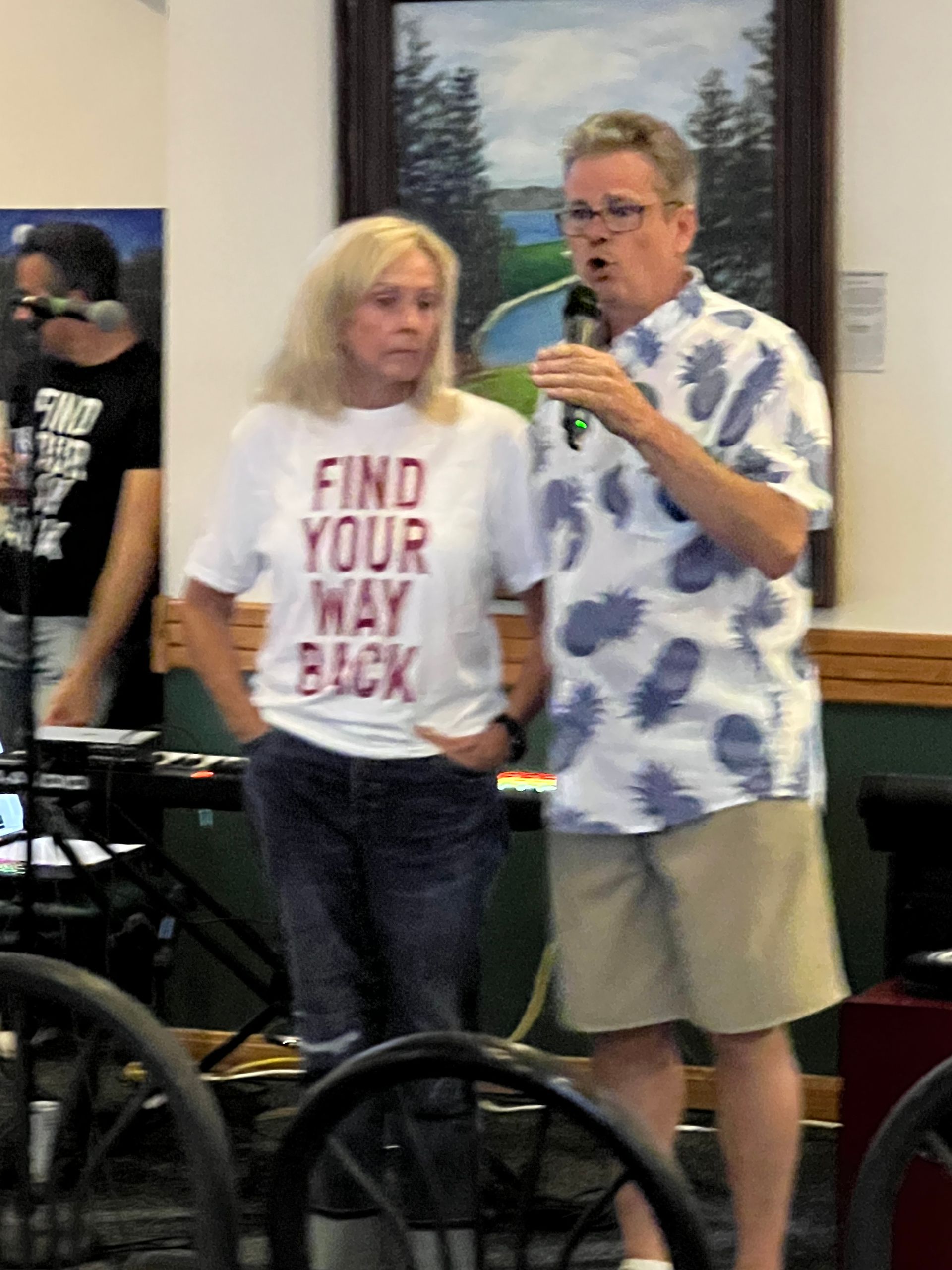 A man speaking into a microphone next to a woman wearing a shirt that says find your way back
