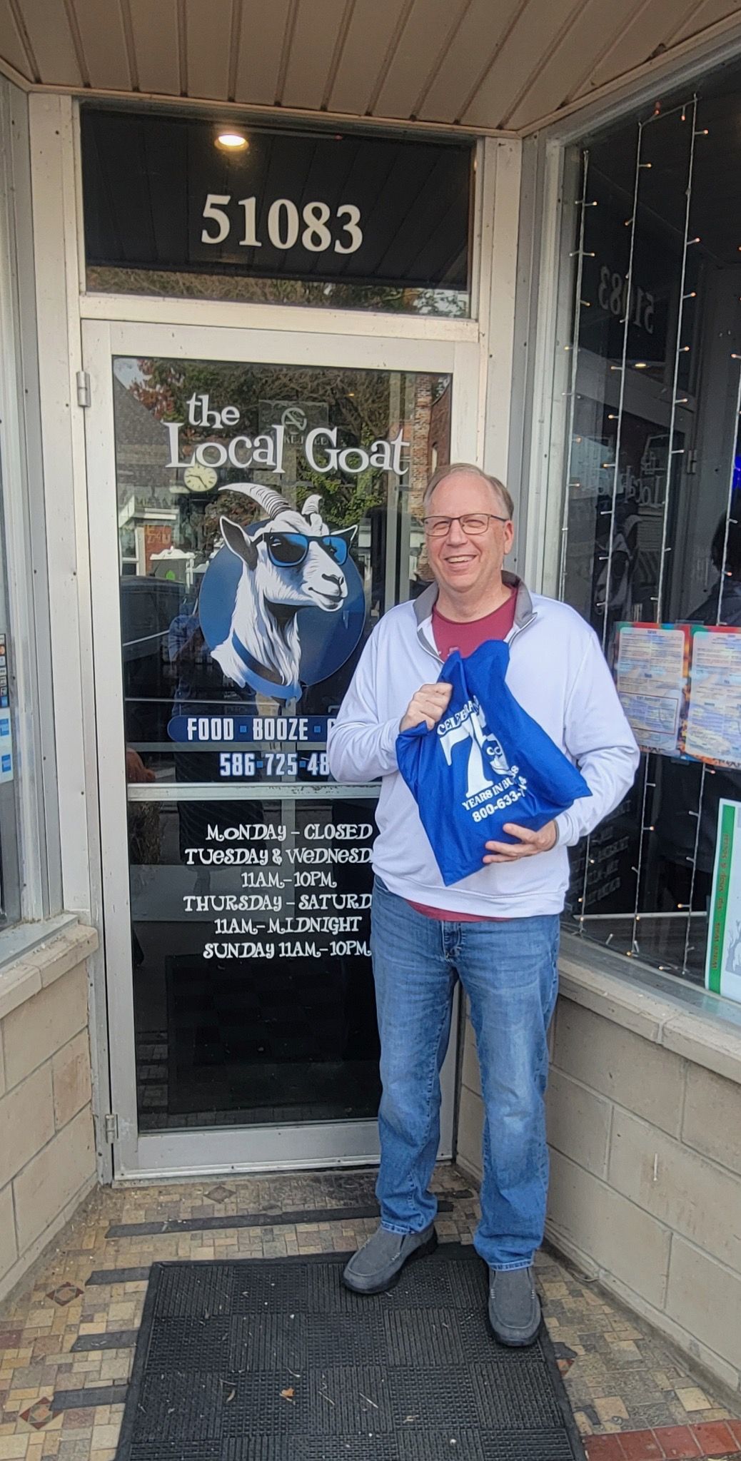 A man is standing in front of a store holding a blue bag.