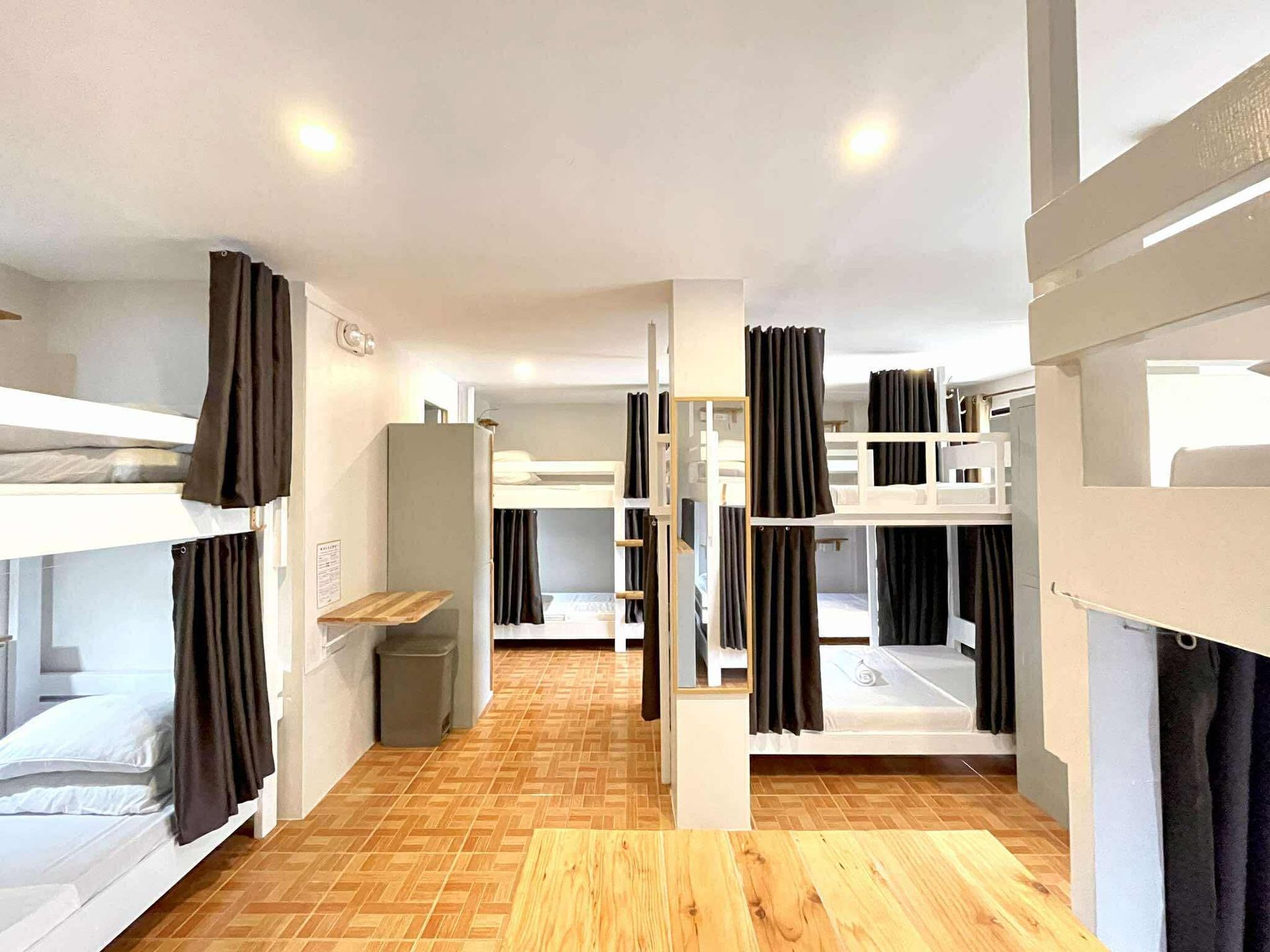 A hostel dormitory with multiple white bunk beds and privacy curtains. Wooden floor and neutral walls.