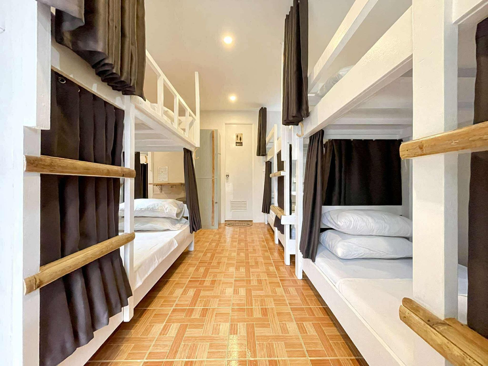 Bunk bed-filled hostel room. White beds, brown curtains. Orange tile floor. Bright overhead lighting. Door at the end.
