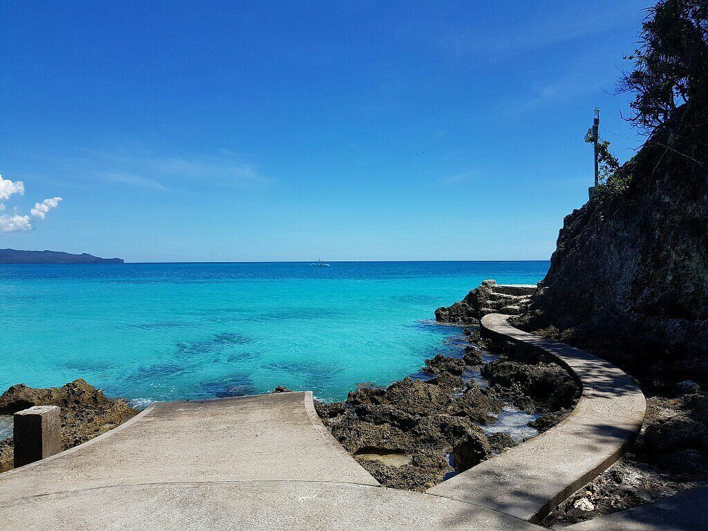 Pathway of concrete slabs leads to turquoise ocean under a bright blue sky.