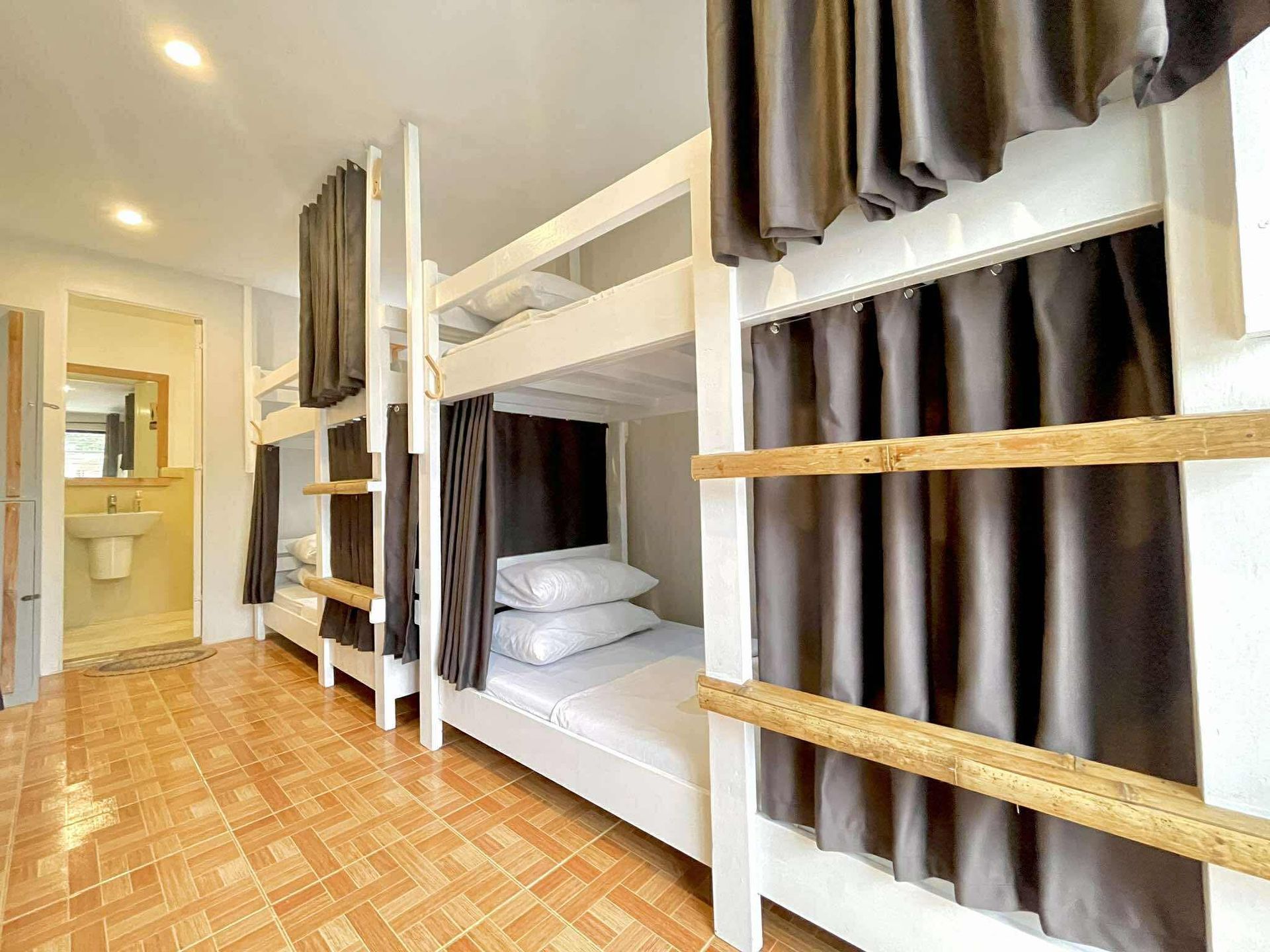 A hostel dorm room with white bunk beds, gray curtains, and a bathroom visible in the background.