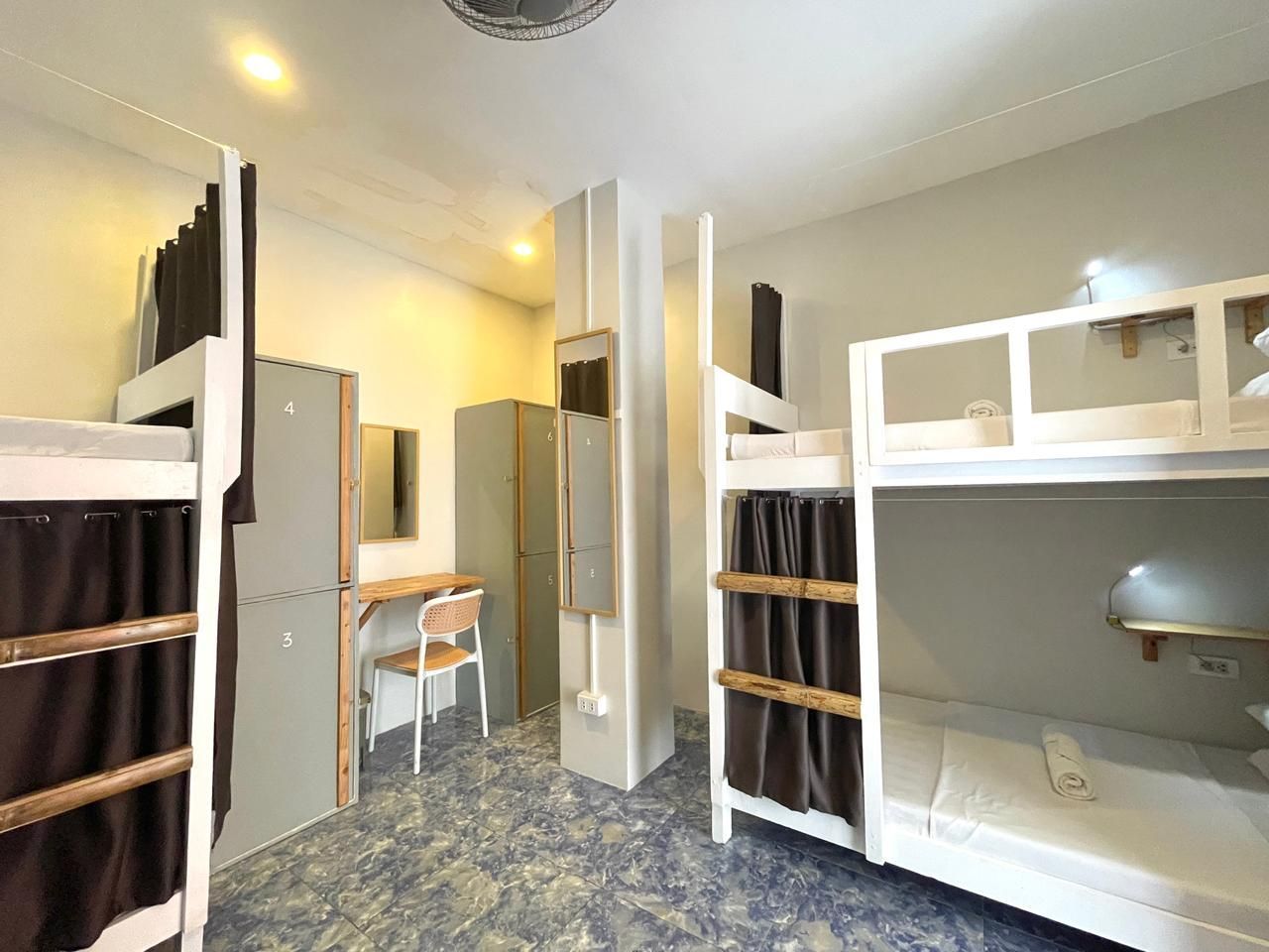 Hostel room with bunk beds, lockers, and a desk. Blue tile floor, white walls.