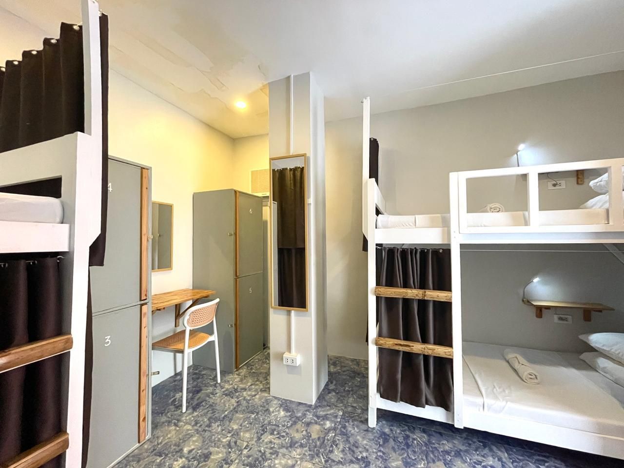 Hostel dorm room with bunk beds, lockers, desk, and mirror. White, gray, and brown tones.
