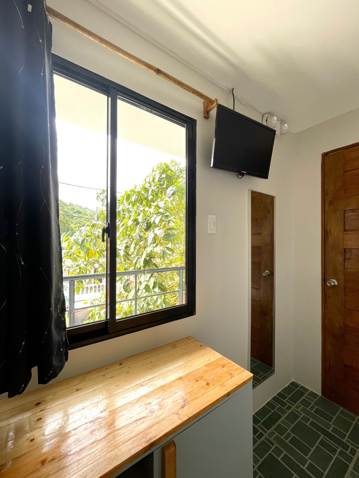 Small room with window, TV, wooden desk, and a door; a balcony is visible outside.