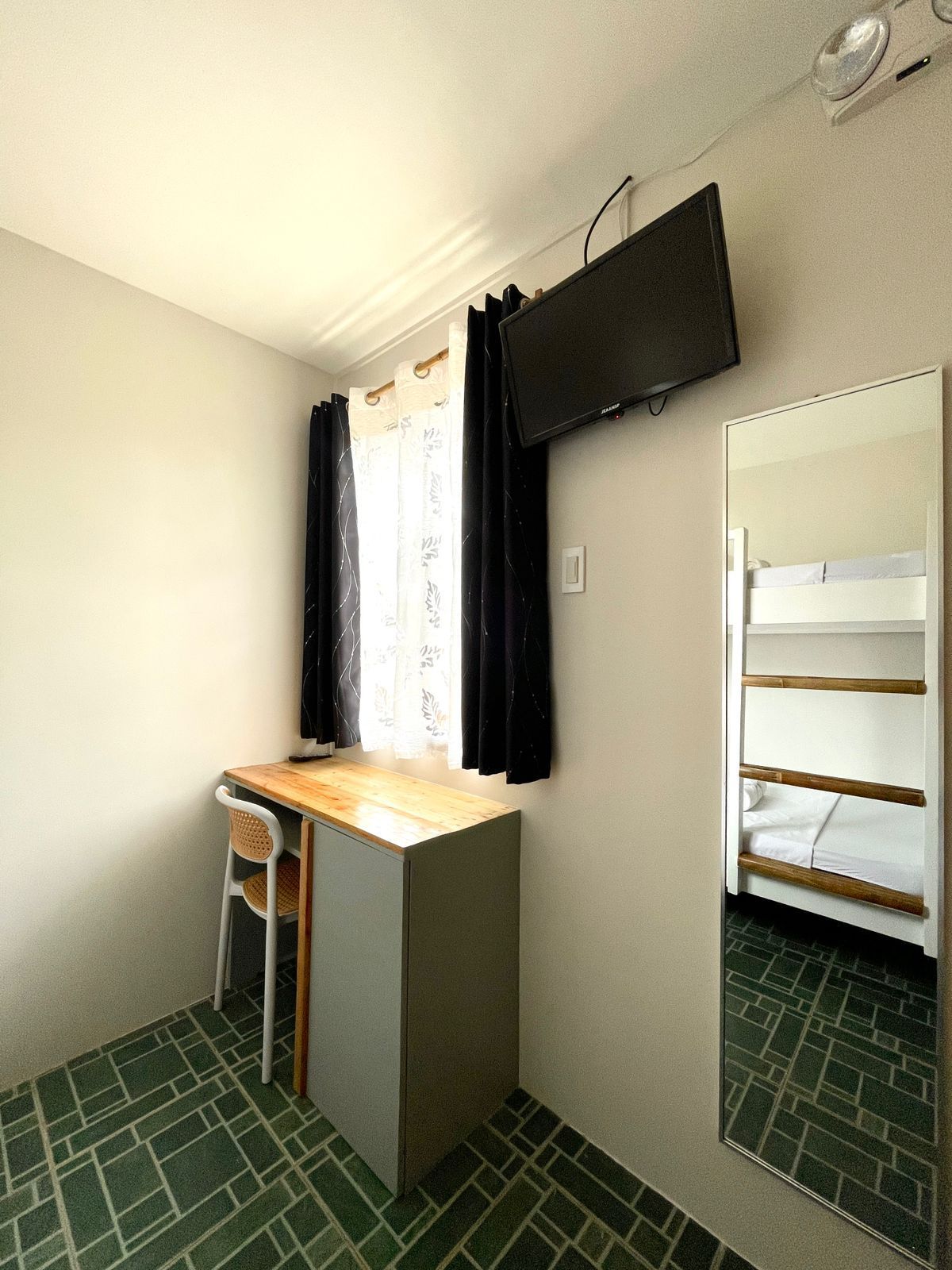 Small hotel room with bunk beds, desk, and mounted TV. Window with dark curtains, green tiled floor.
