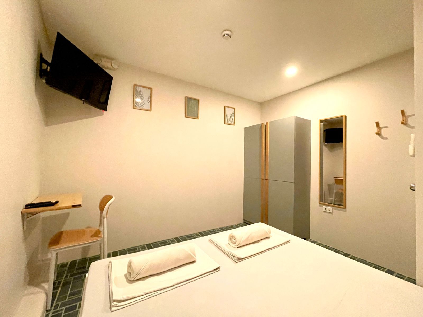 Small hotel room with a bed, TV, desk, wardrobe, and mirror. White walls and grey accents.
