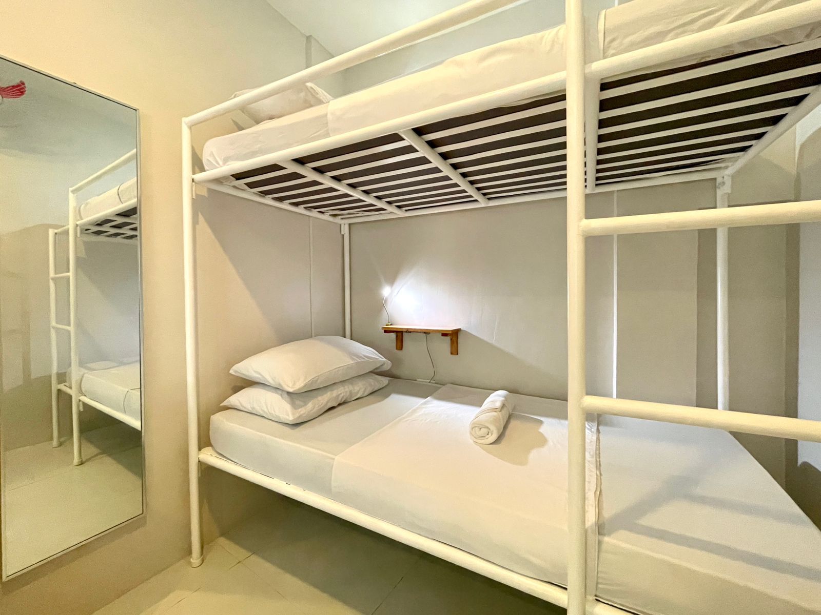 Bunk beds in a hostel room. White metal frame, white linens.  Mirror on the wall reflects the beds.