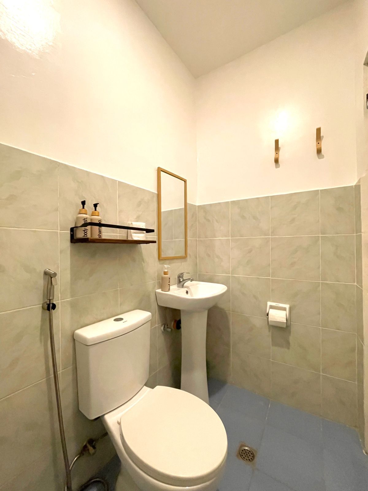 Small bathroom with toilet, pedestal sink, mirror, shelf, and blue floor. Light-colored tile walls.