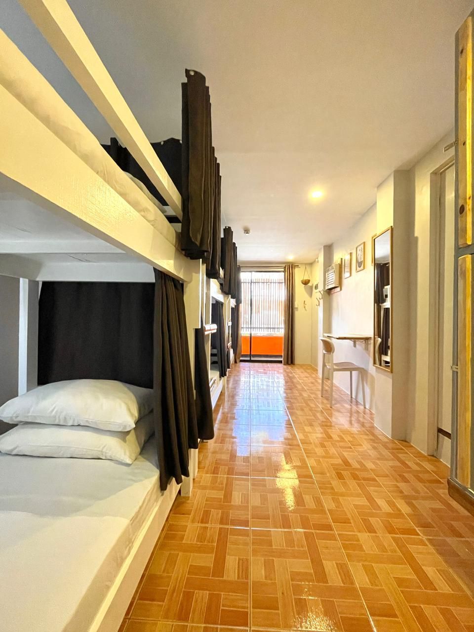 Bunk beds line a narrow hostel hallway with tiled floors. Brown curtains offer privacy.