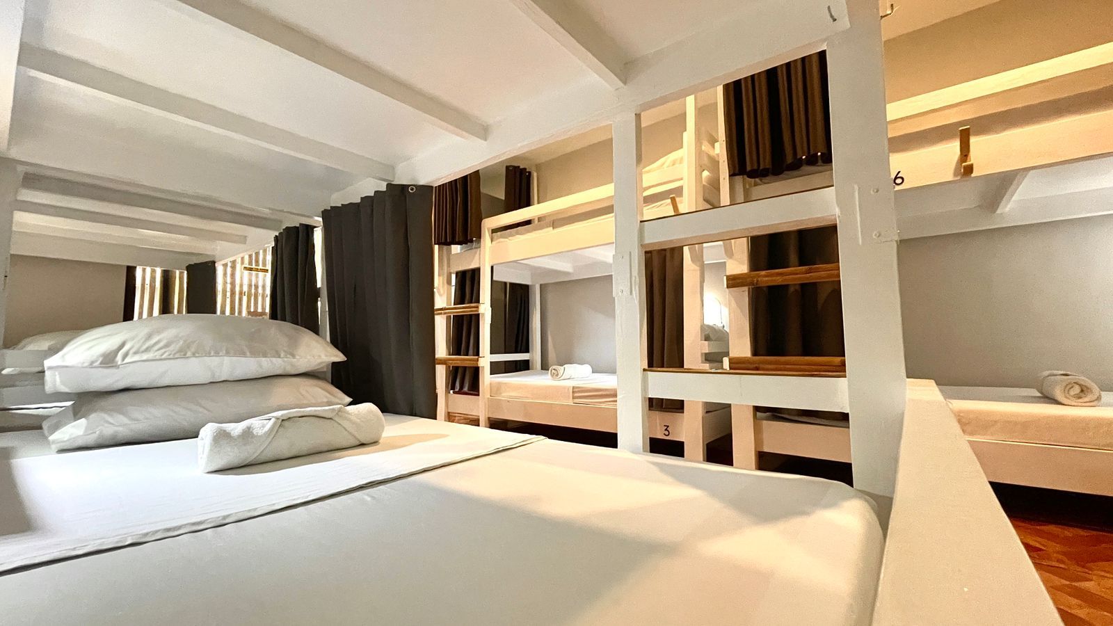 Bunk beds in a well-lit hostel room. White sheets and pillows, wooden frames, and gray curtains.