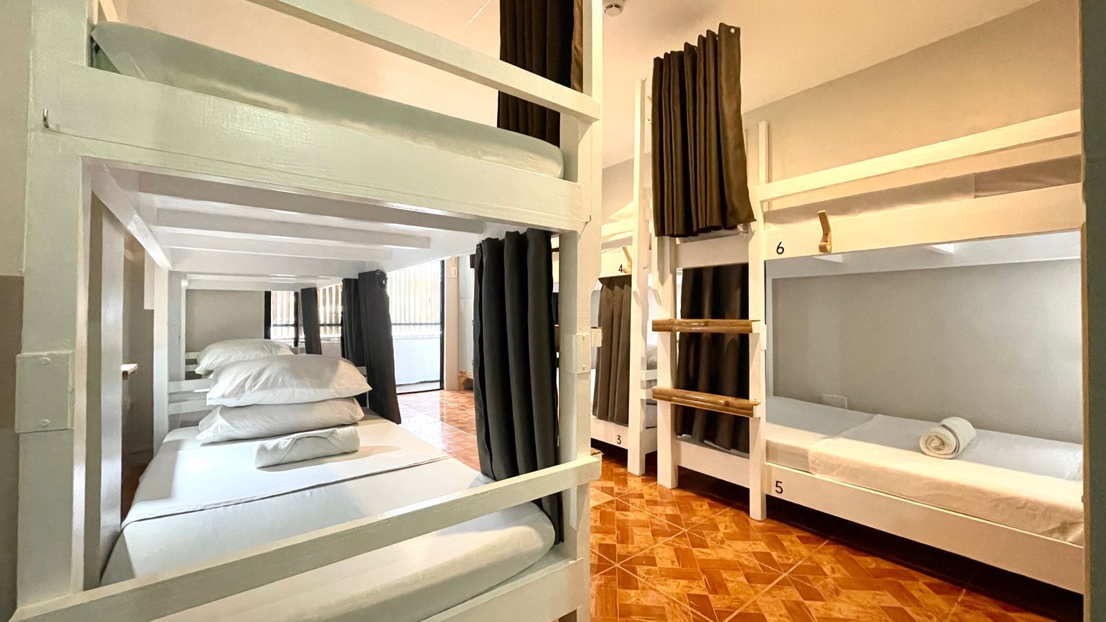 Hostel room with multiple white bunk beds, gray curtains, tiled floor.