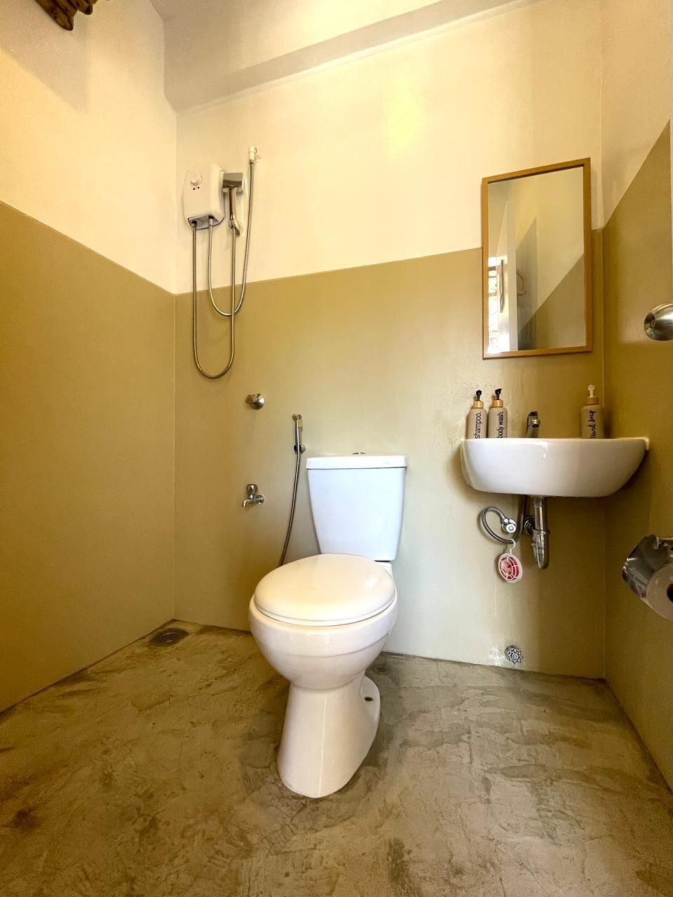 Bathroom with toilet, sink, mirror, and showerhead. Beige and white walls, concrete floor.