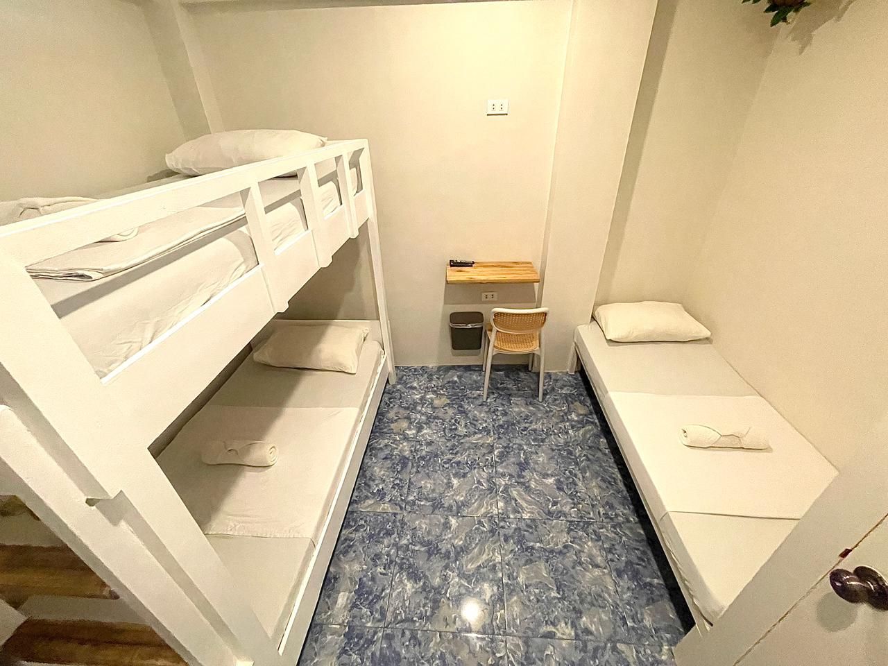 A small, white-walled room with a bunk bed, single bed, desk, and blue tiled floor.