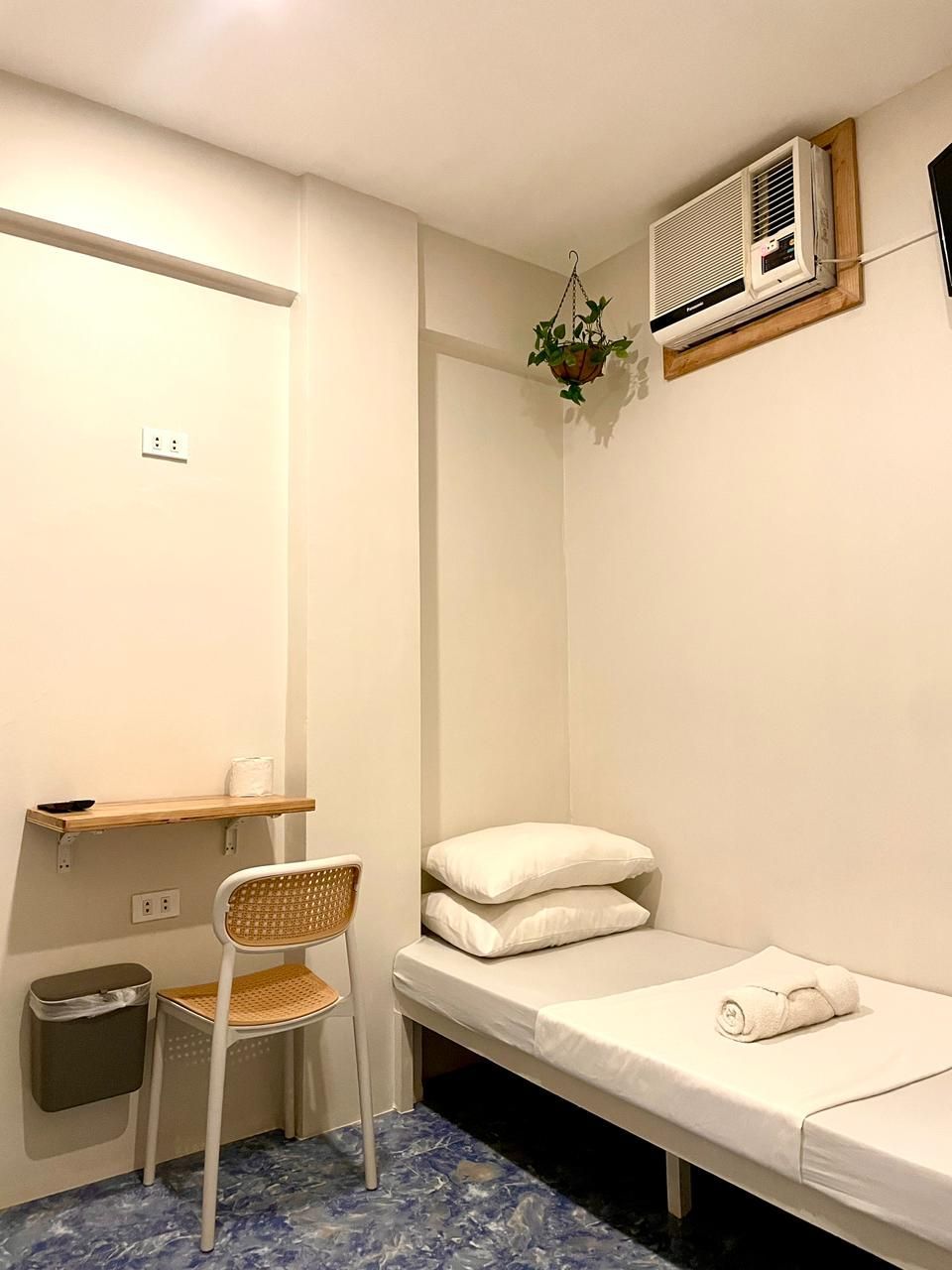 Small, white-walled room with a bed, air conditioner, and floating shelf. Blue floor, hanging plant.