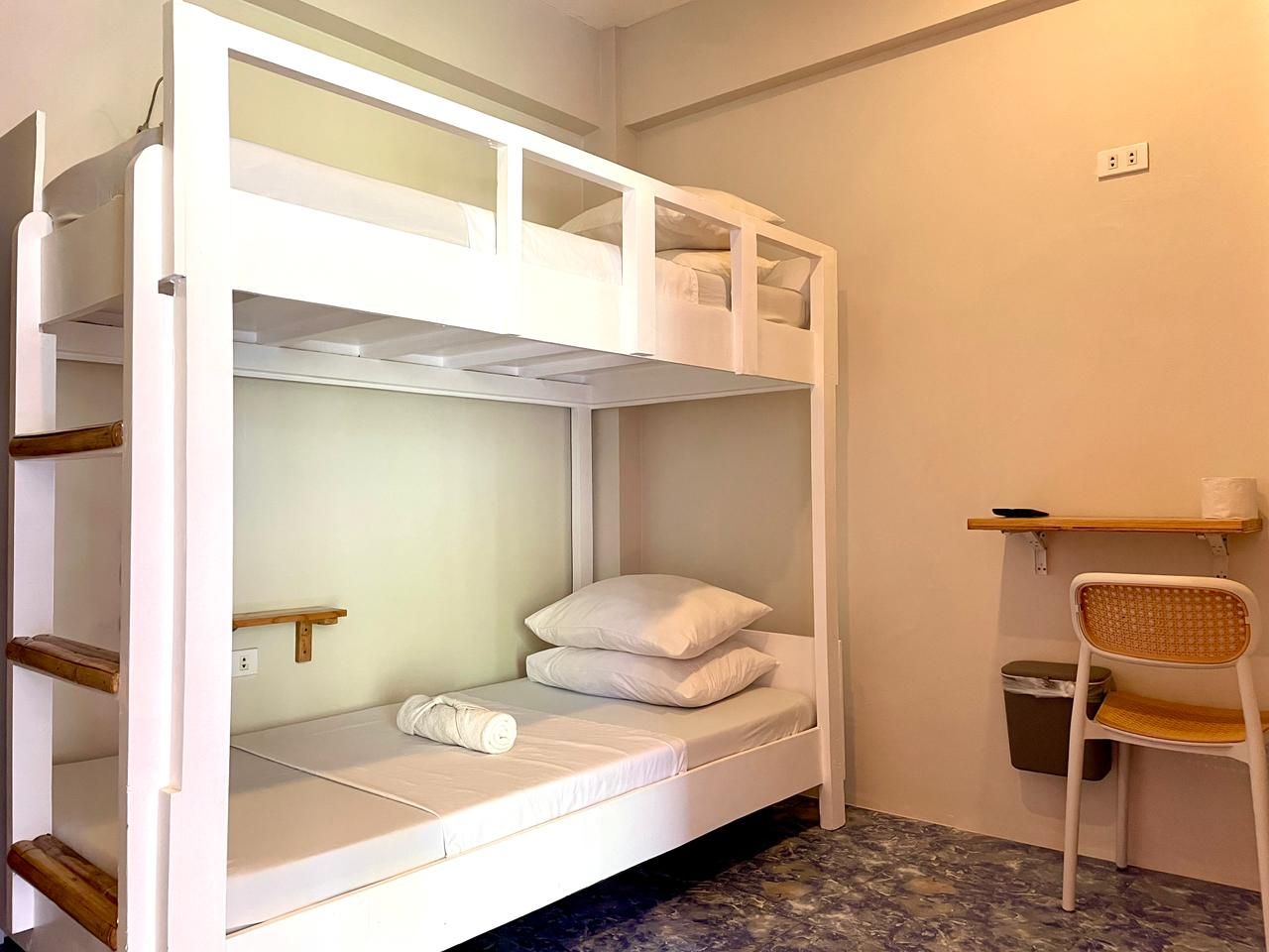 Bunk beds in a light-colored room with simple decor. A chair and small shelves are visible.