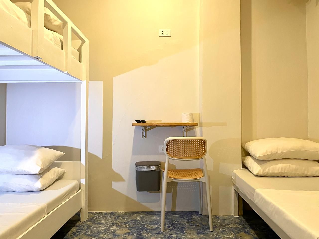 Bunk beds and single beds in a room. A small desk, chair, and trash can are in between. Beige walls.