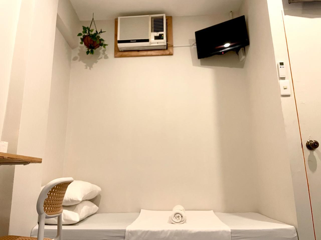 Small, white room with bed, hanging plants, air conditioner, and wall-mounted TV.