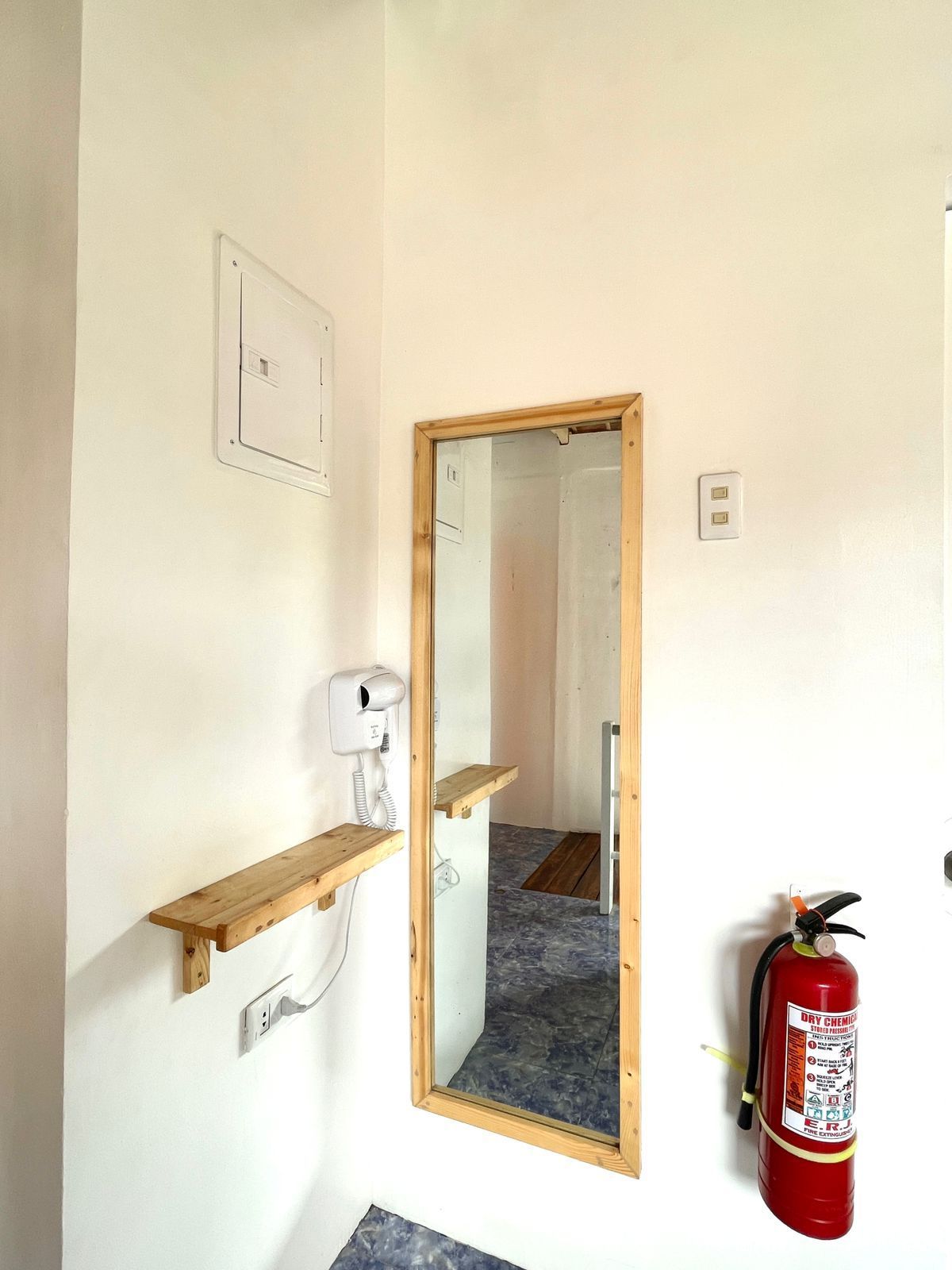 Corner of a room with mirror, shelf, hairdryer, fire extinguisher, and electrical panel.