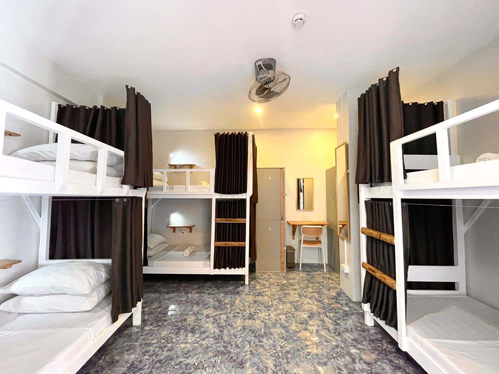 Hostel room with multiple white bunk beds and privacy curtains; gray lockers and a desk.