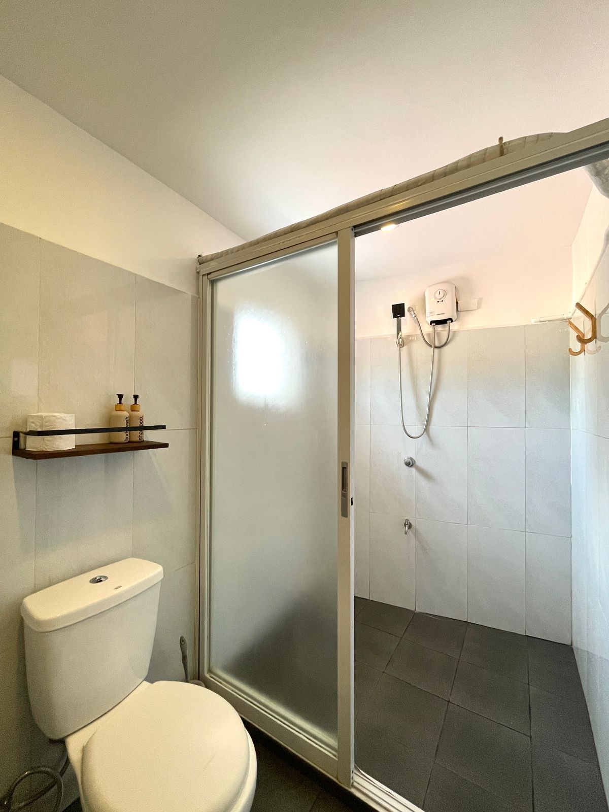 A bathroom with a toilet, shower, and shelf with toiletries. The walls are tiled.