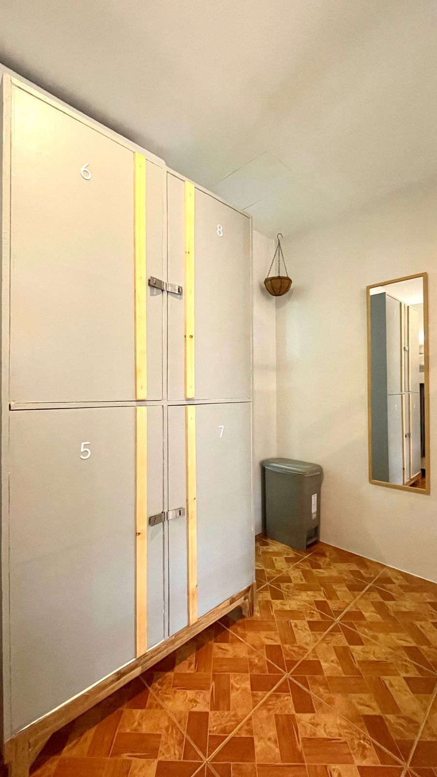 Tall grey lockers, brown tiled floor, mirror, small trash can, hanging plant.