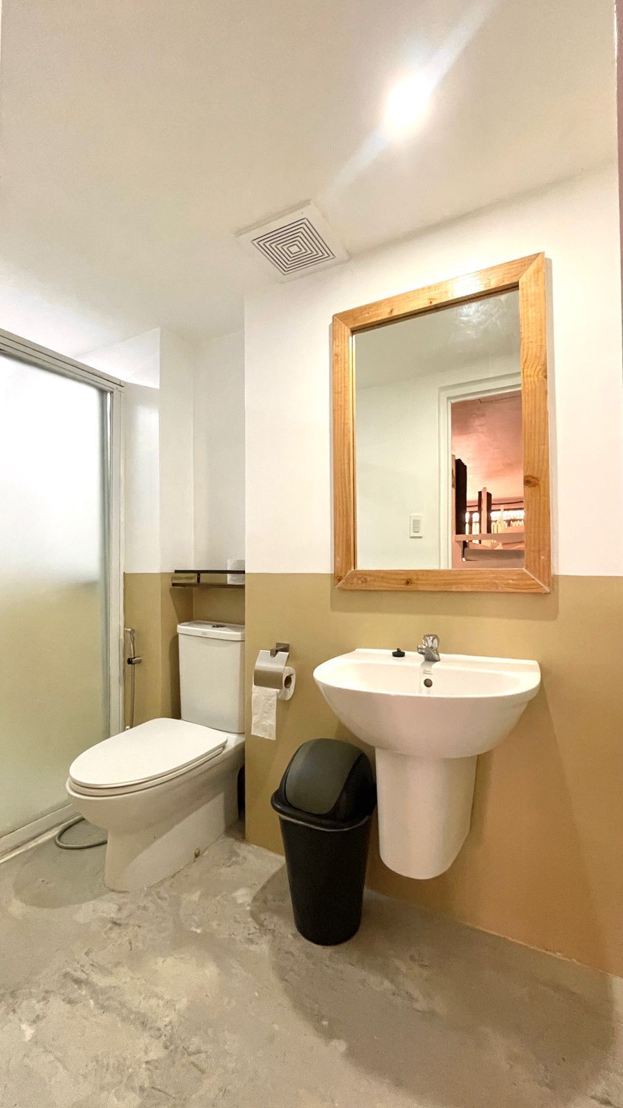 Bathroom with toilet, sink, mirror, and trash can. Tan and white walls, gray floor.