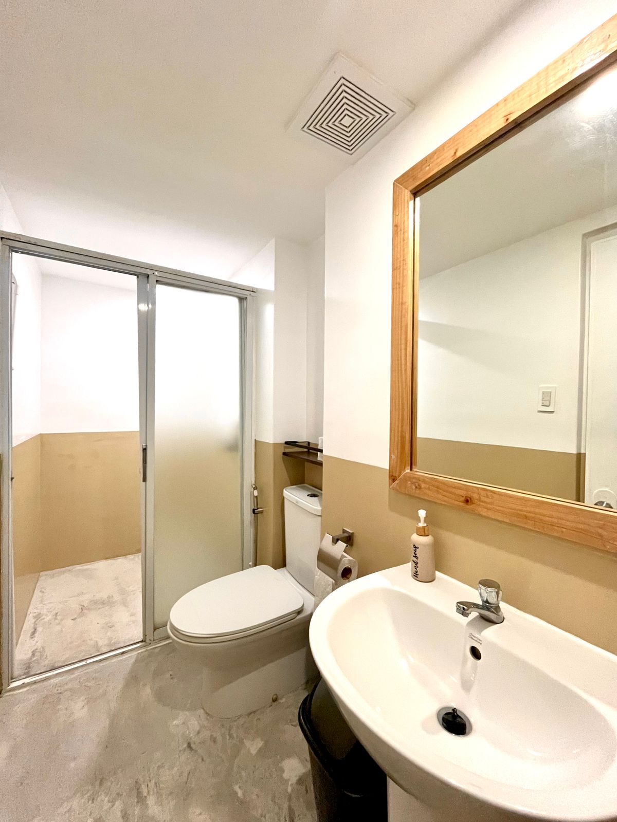 Bathroom with toilet, sink, mirror, and shower. Tan and white walls, concrete floor.