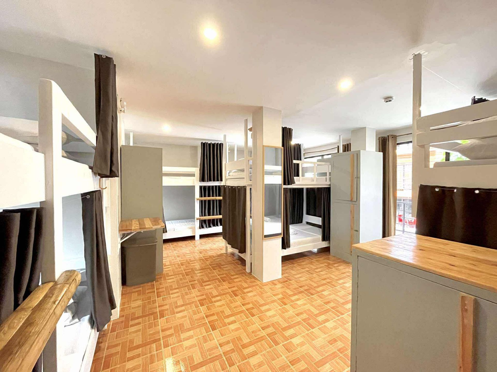 Hostel room with multiple white bunk beds and privacy curtains; wood floor.