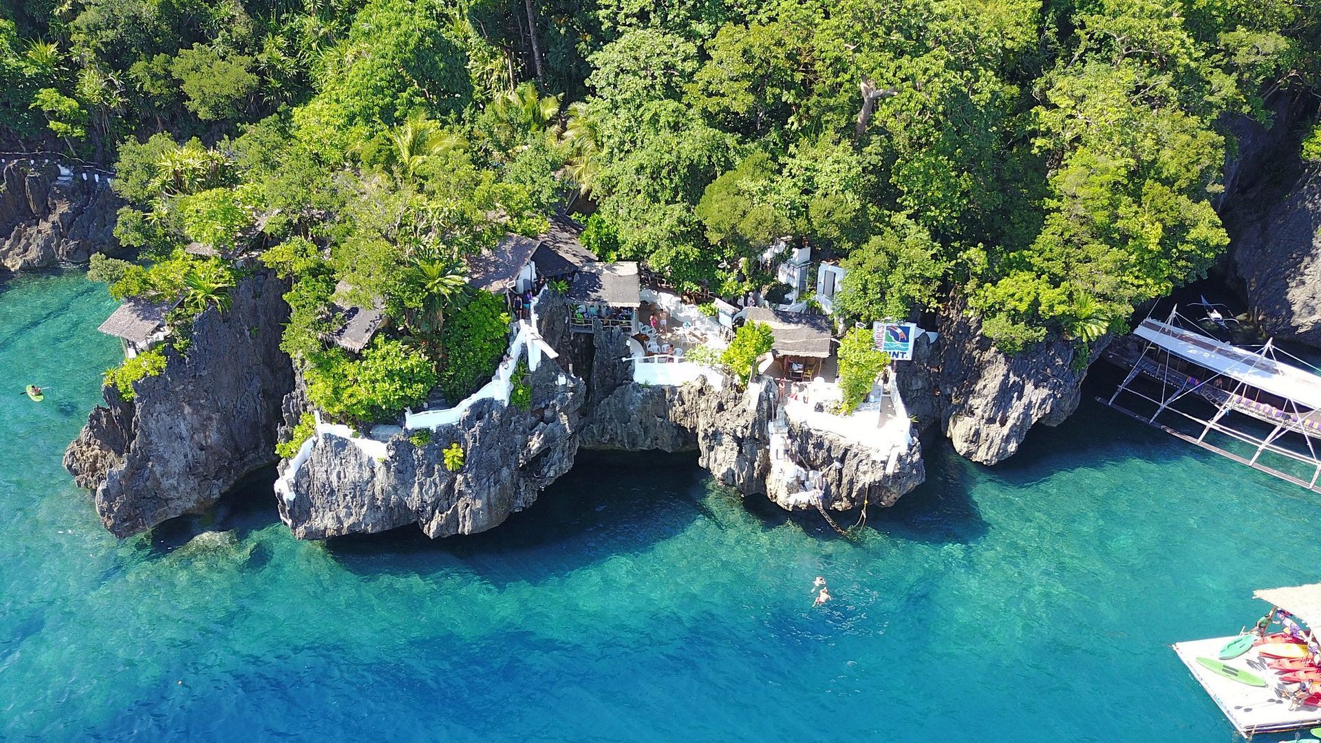 Aerial view of a rocky island with lush greenery, buildings, and clear turquoise water.
