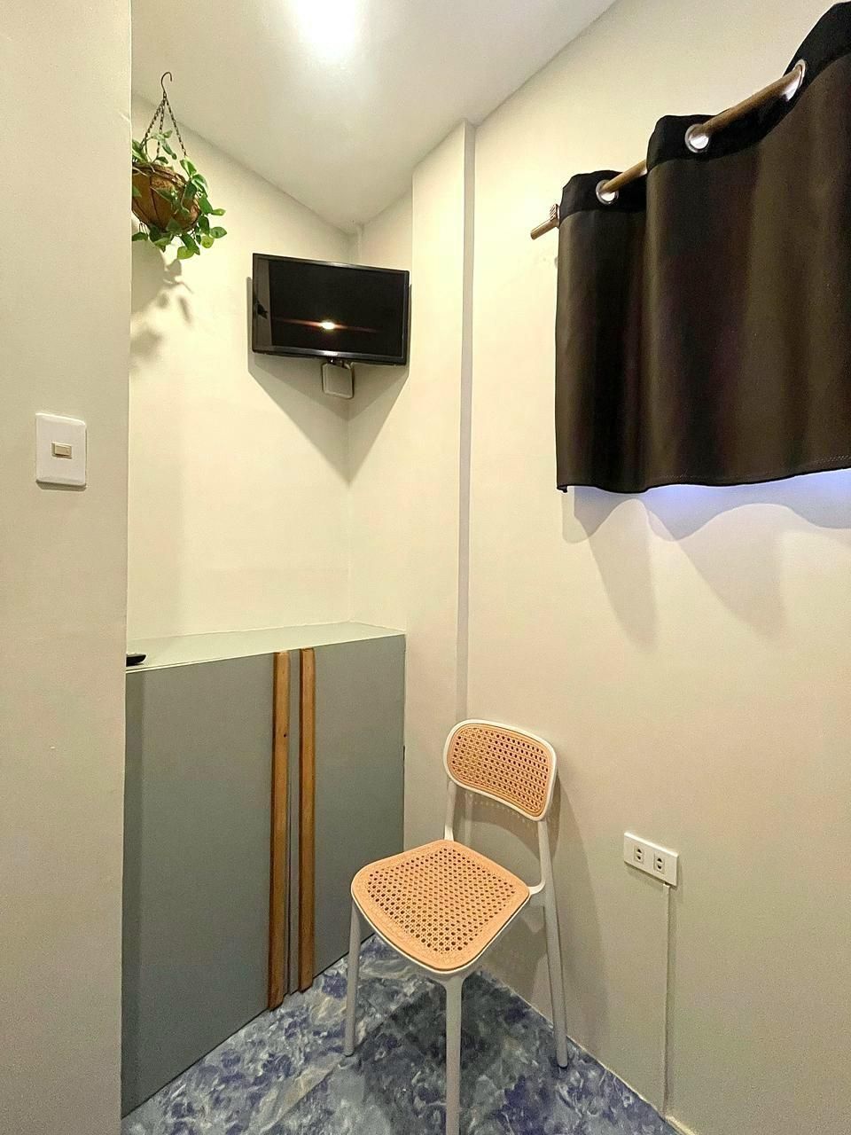 Small room with TV, chair, and cabinet. Blue tiled floor, brown curtain, hanging plant. Light-colored walls.