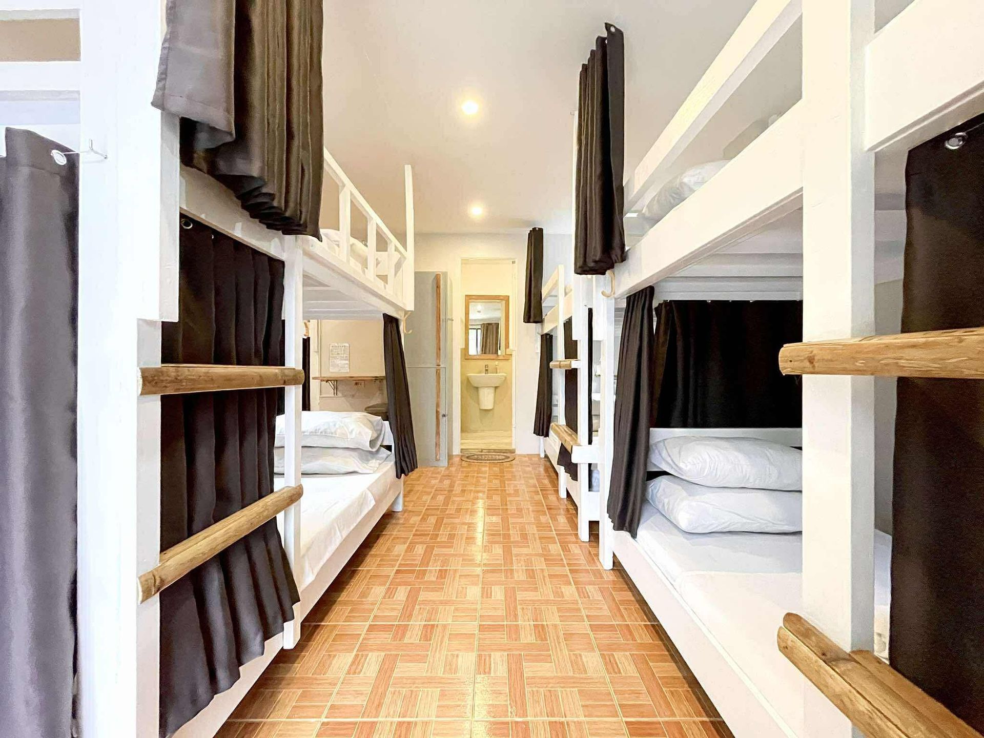 Hostel dorm room with white bunk beds, gray curtains, and tiled floor. A sink and mirror are visible in the background.