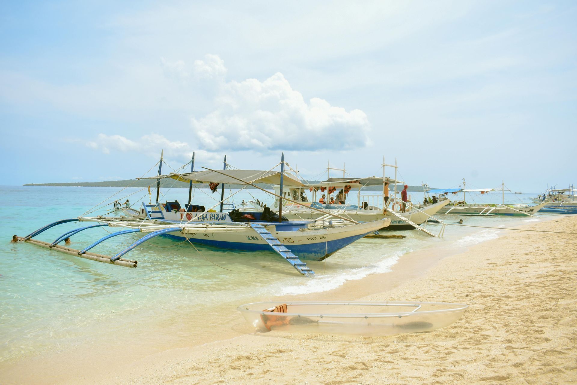 Boats docked on a sandy beach with clear, turquoise water under a blue sky.