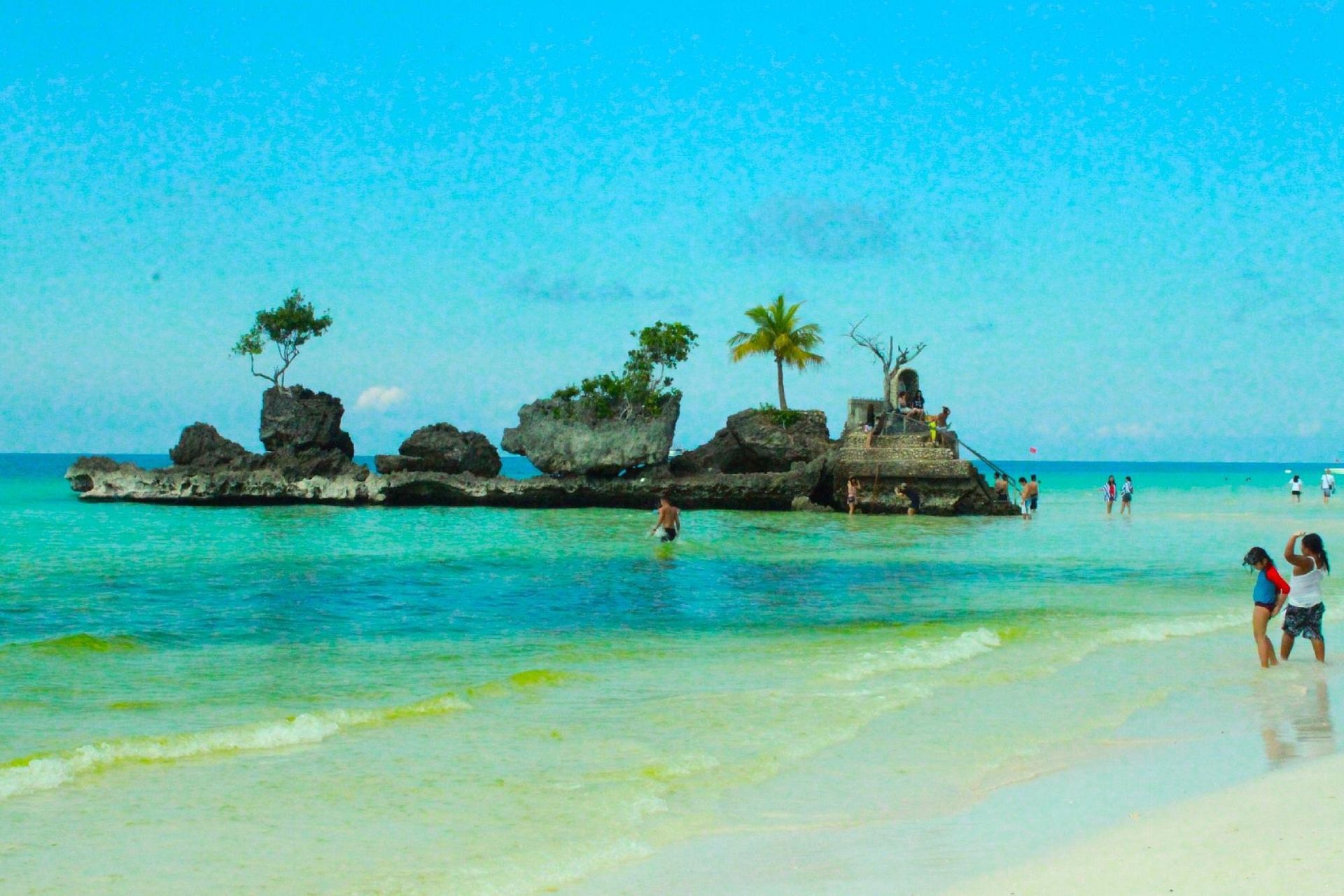 Rock formations with trees in turquoise water, people on the beach and in the water, under a blue sky.