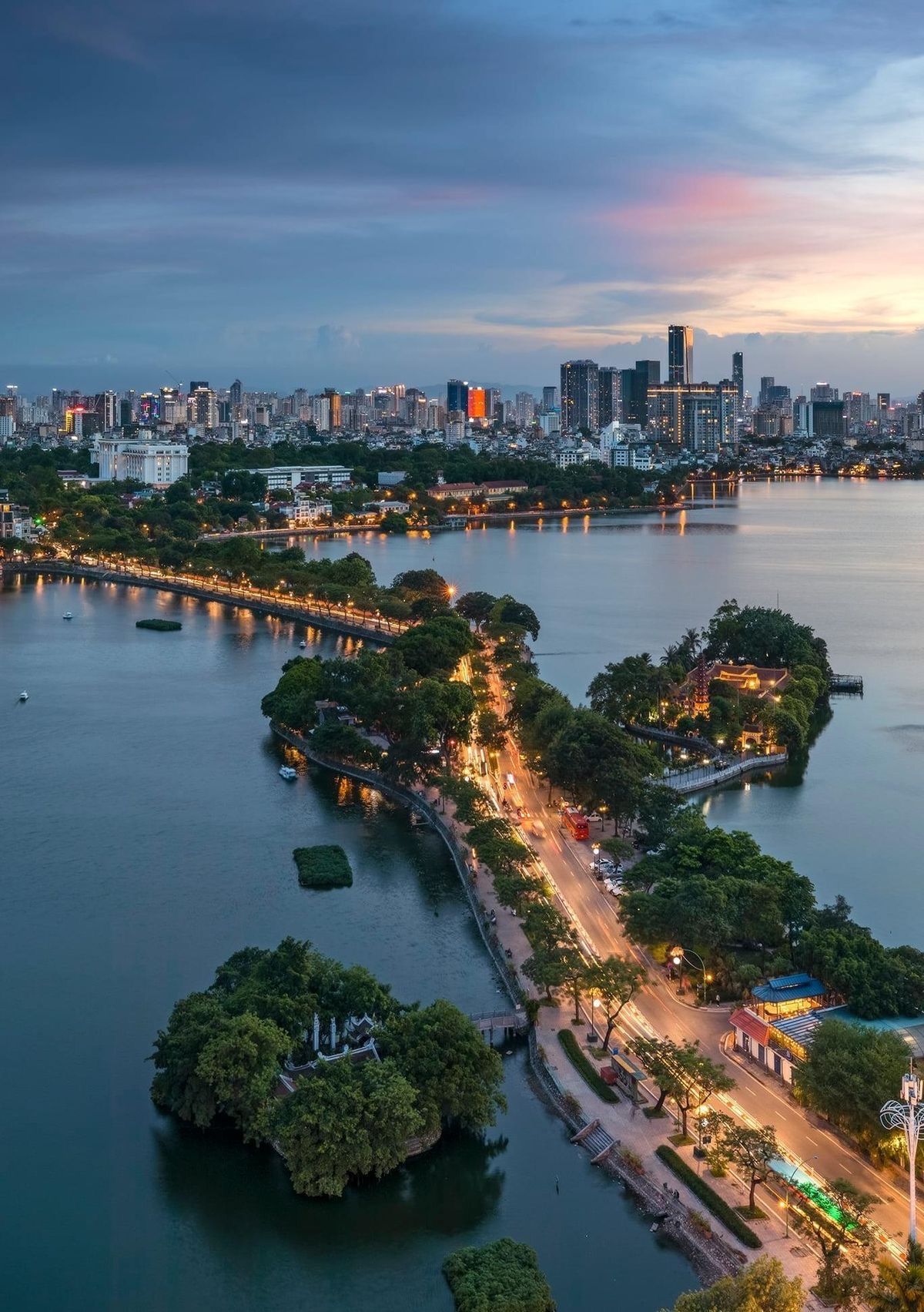 Cityscape at dusk, a road with lights curving across a lake towards buildings in the distance, blue and pink sky.
