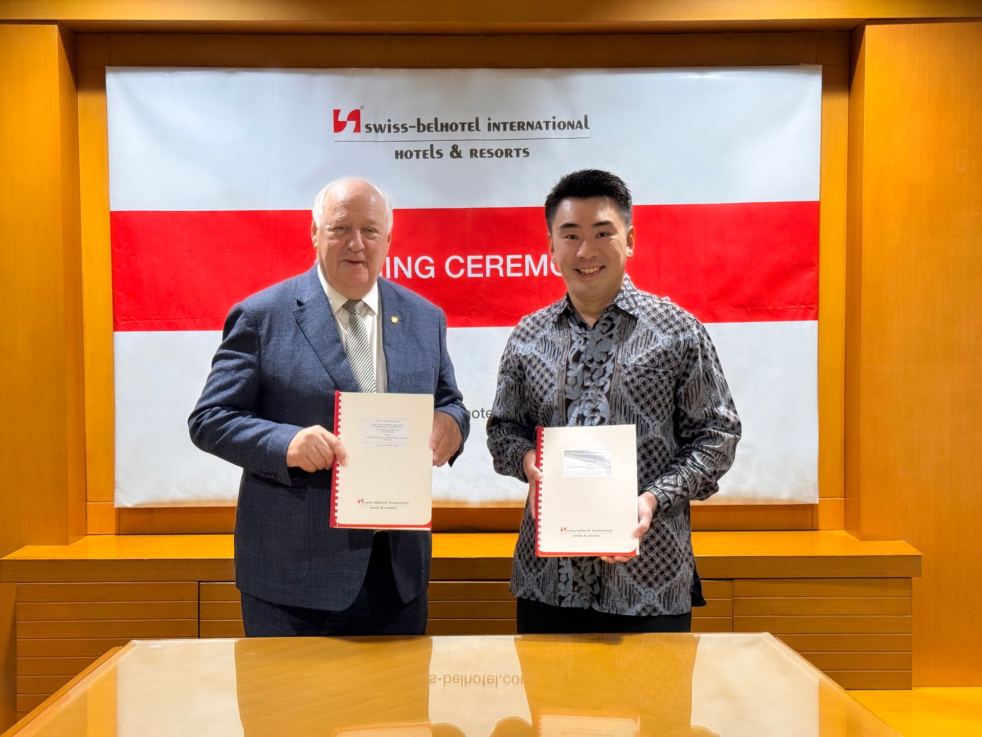Two men holding documents, in front of a banner. One wears a suit, the other a patterned shirt. They are in a wood-paneled room.