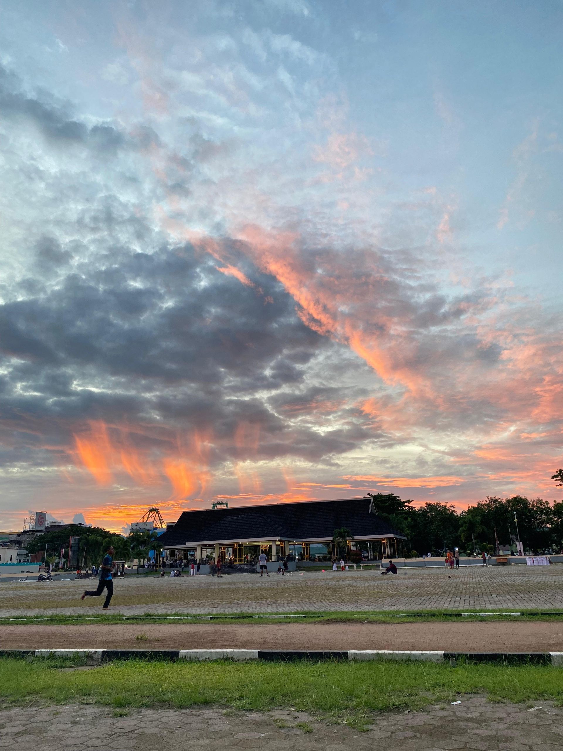 Sunset sky over a park; person running. Orange and gray clouds, green grass, building.