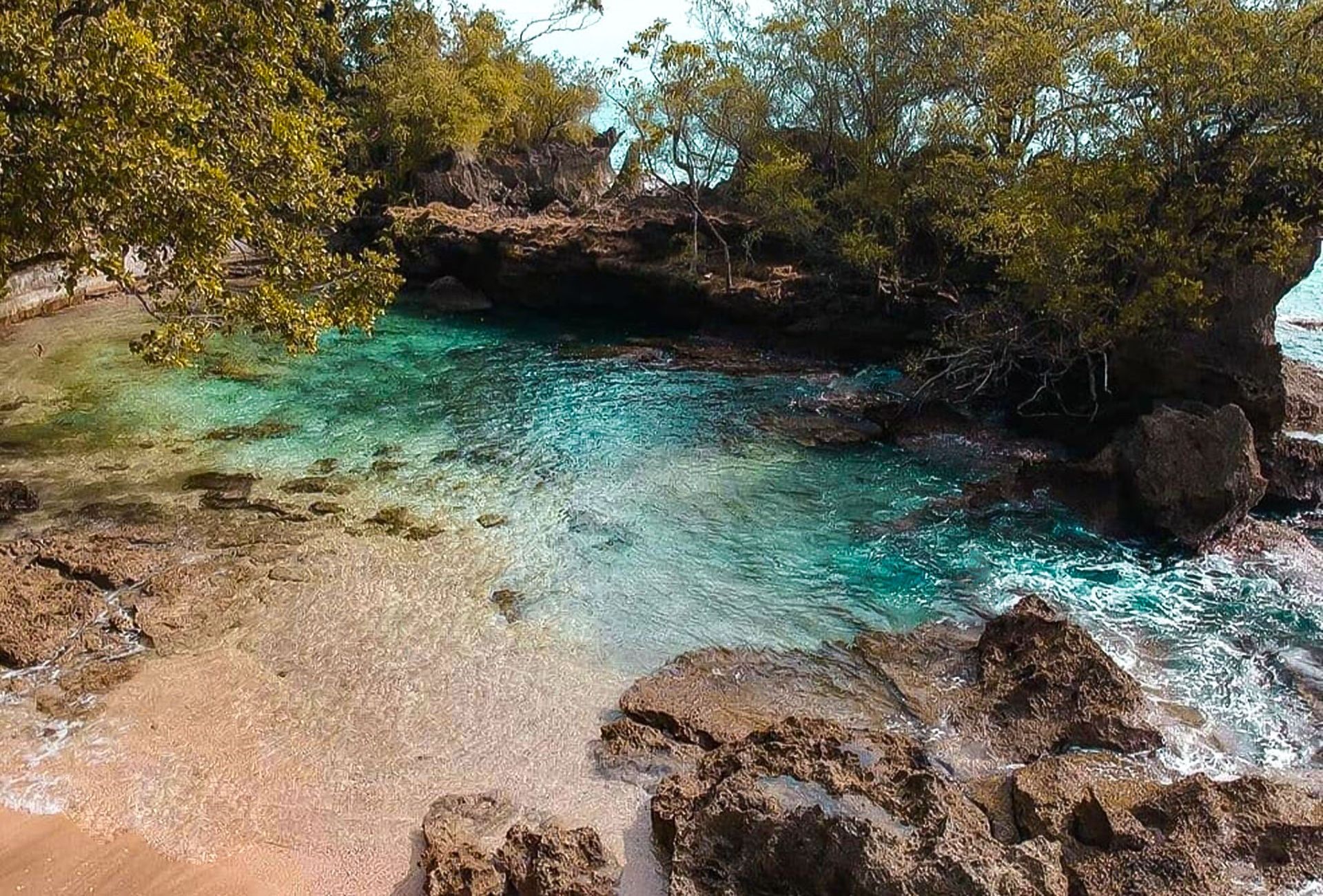 Small turquoise lagoon surrounded by rocks and trees, with sandy beach.