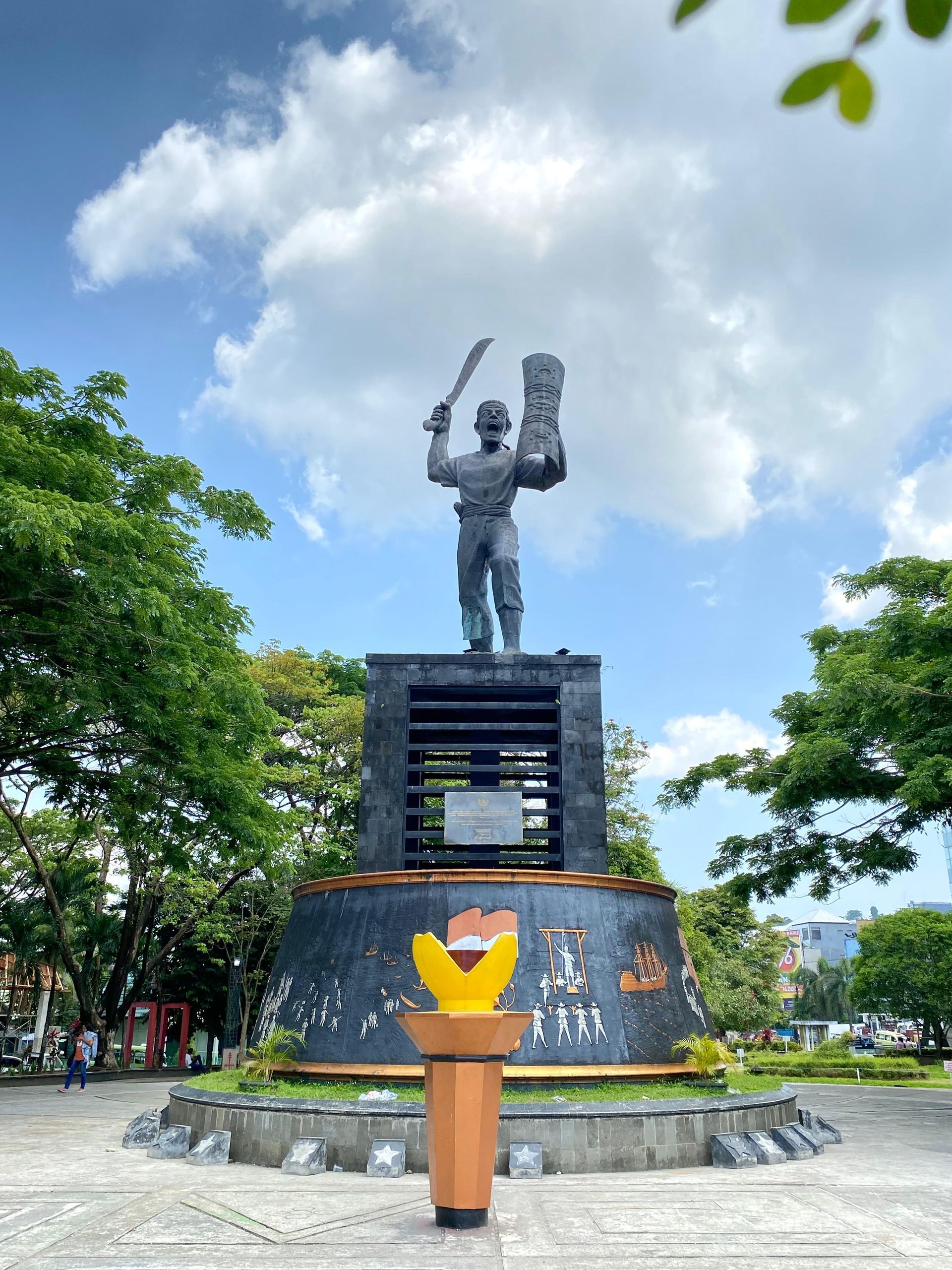 Statue of a man holding a sword and a book on a pedestal, set in a park. Cloudy sky, trees.