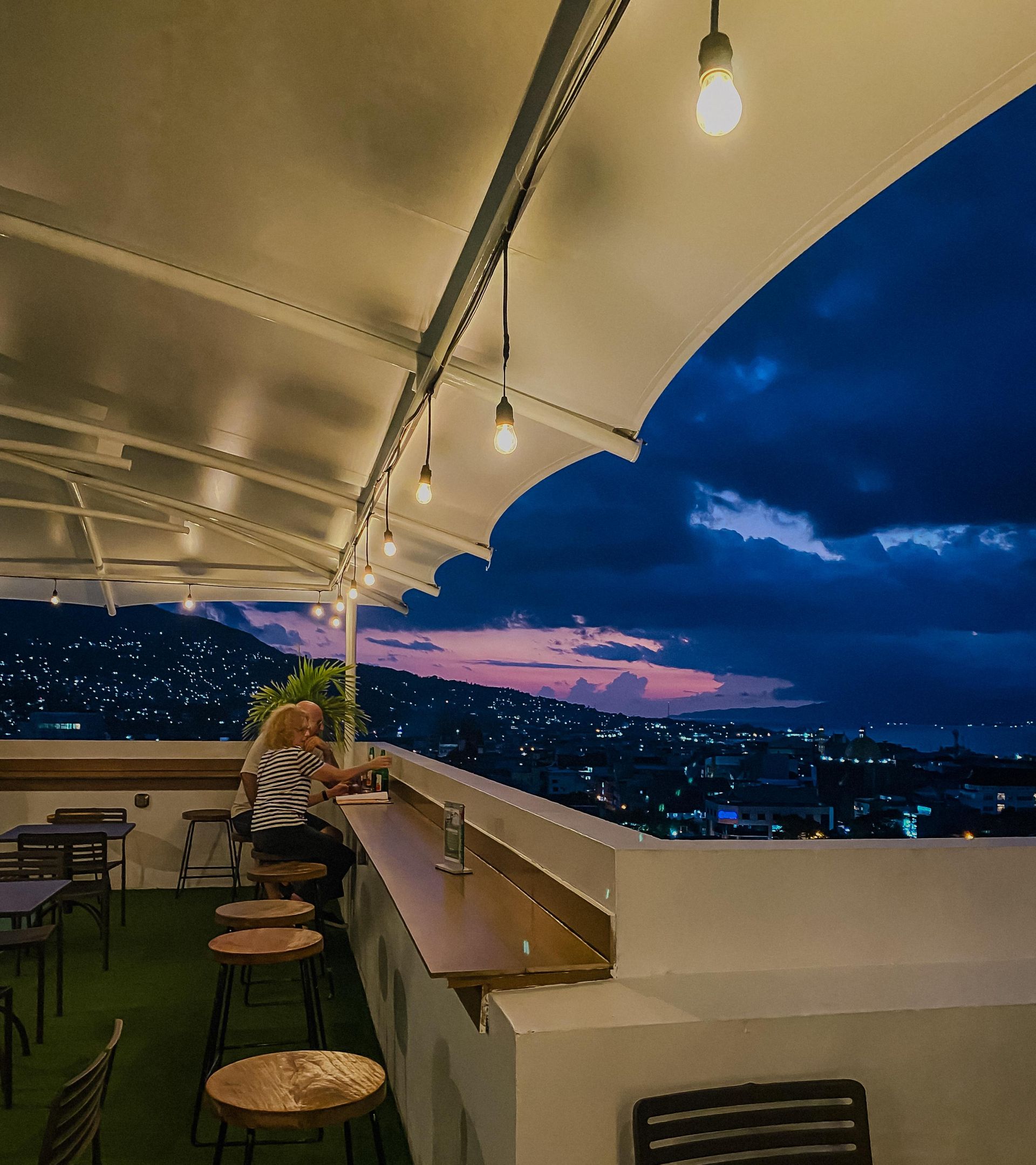 Rooftop bar at dusk with city view; woman seated at the bar under string lights.