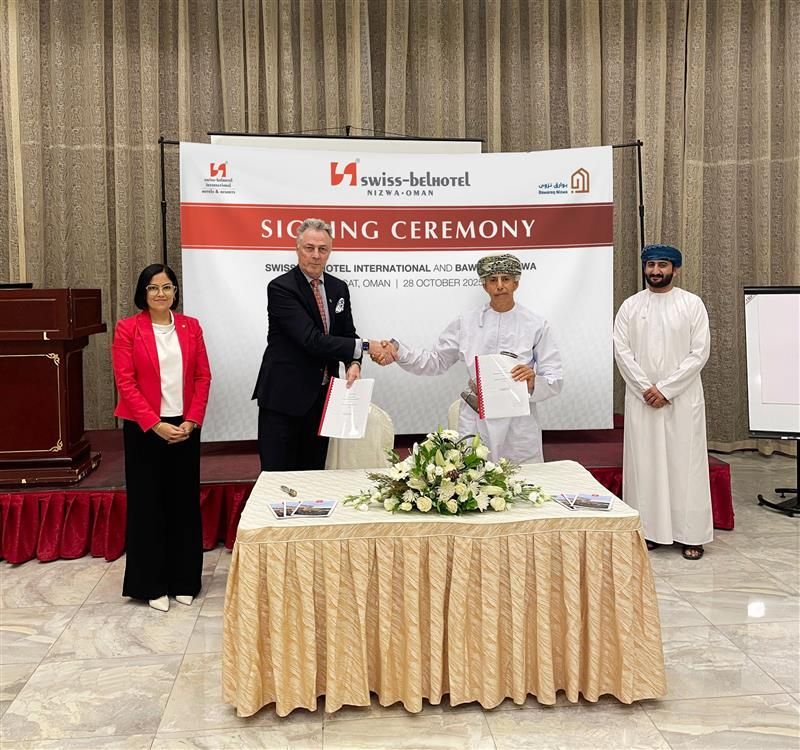 People at a signing ceremony in a hotel. Two men shake hands over a table with documents. Swiss-Belhotel banner in background.